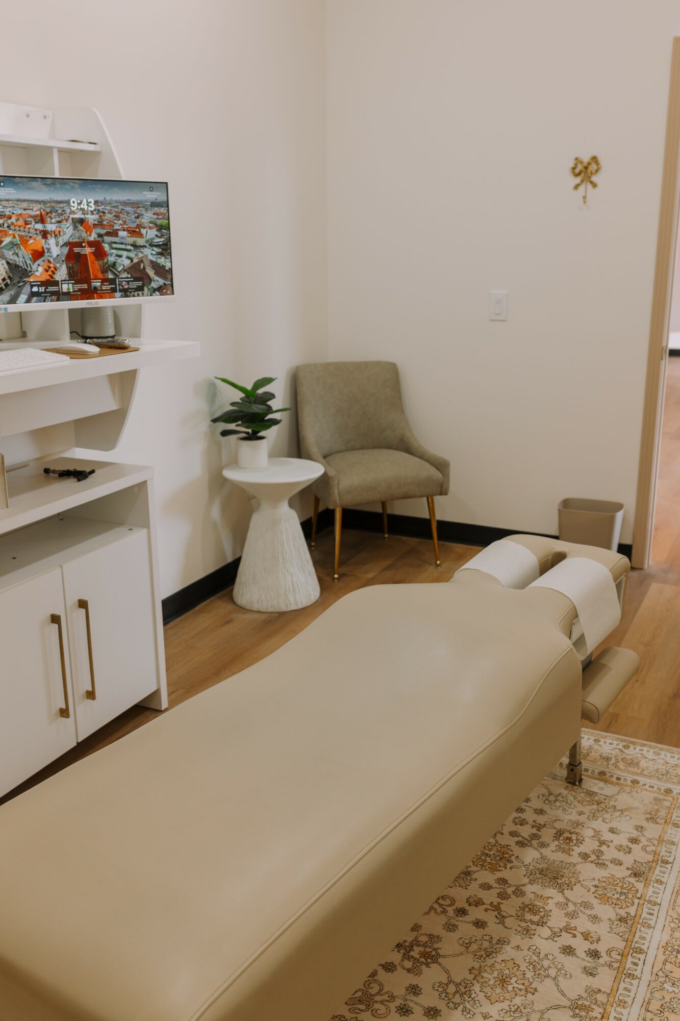 Upright view of a room with a beige bed, a green armchair, a small white table with a plant, and a TV on a white shelf.
