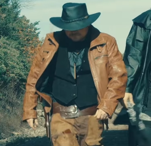 Person wearing a black hat, brown leather jacket, and black clothing outdoors, walking on a dirt path.