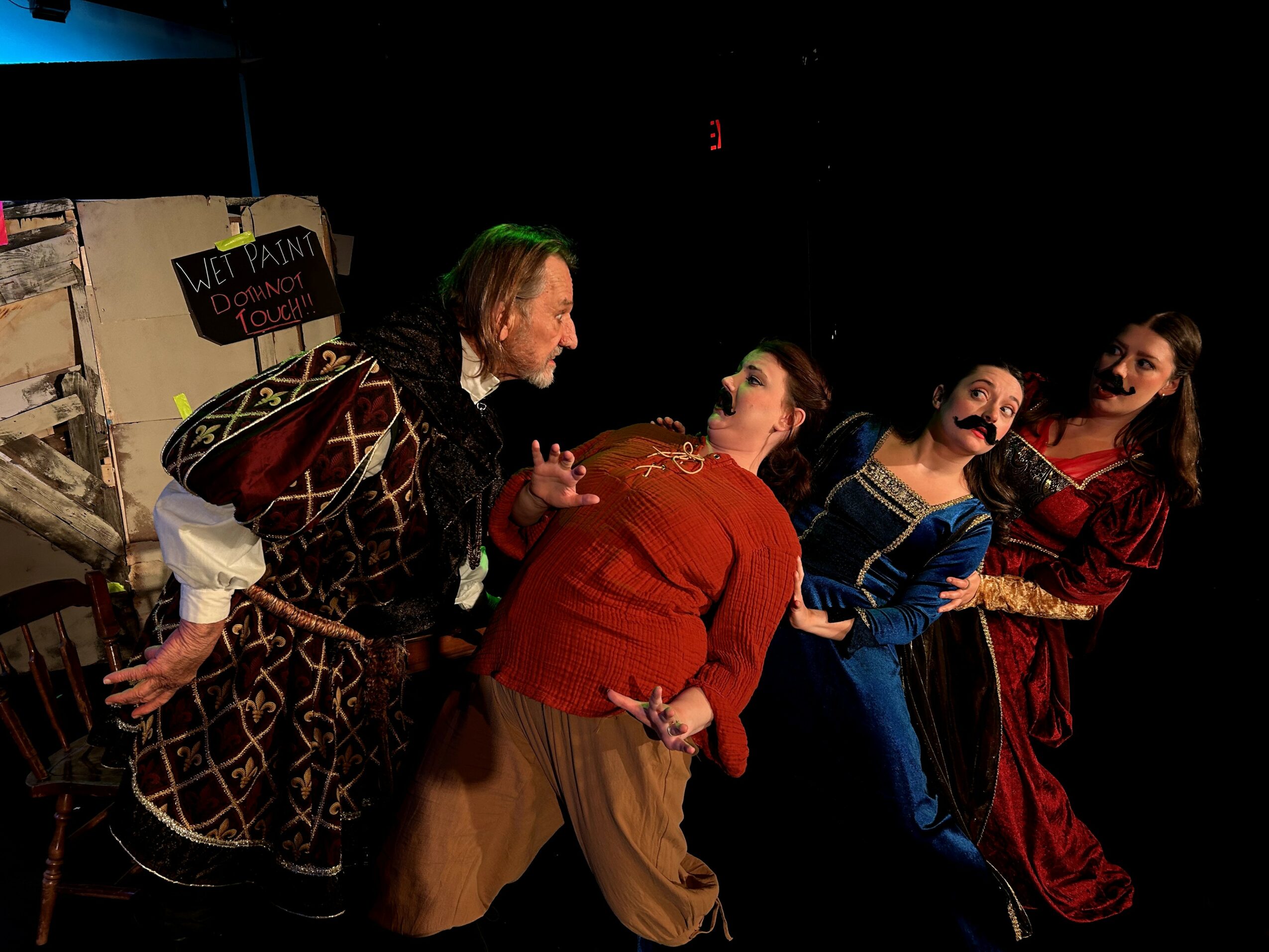 Four people in costumes engaging in a theatrical scene, with a cardboard box and props in the background.