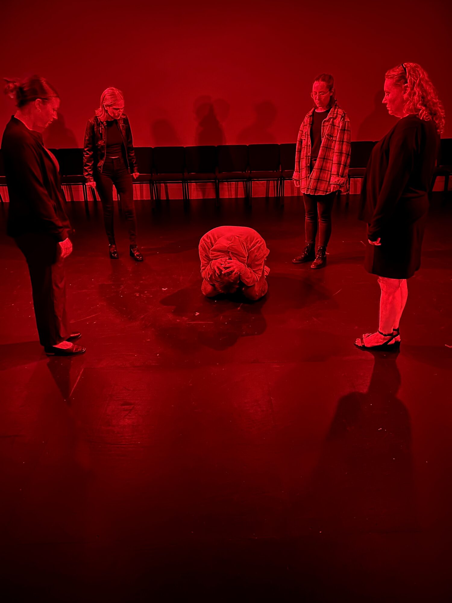 Five people stand in a circle around a person kneeling on the floor, all in a dimly lit room with red lighting.