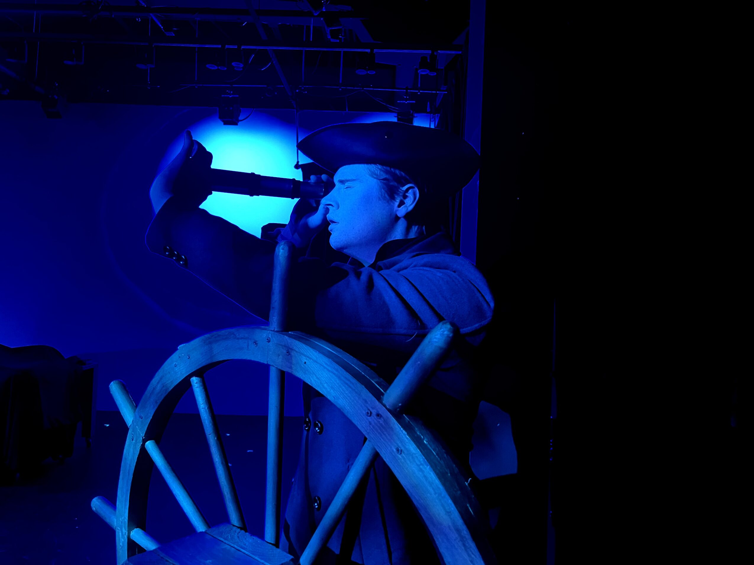 Person wearing a cap, holding a microphone, with a large wooden wheel in front, illuminated by blue light.