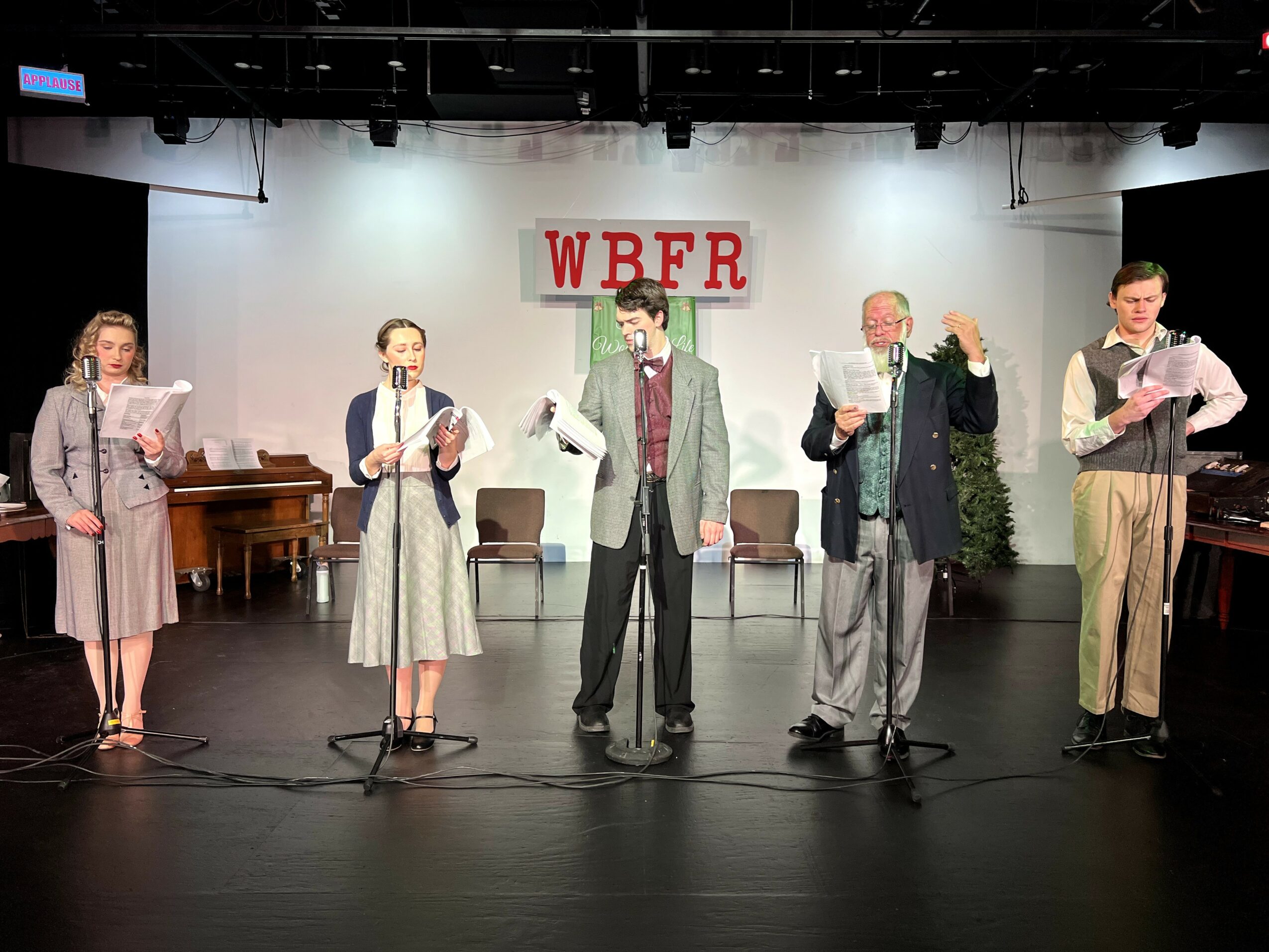 Five people stand on stage reading from scripts, with a WBFR sign behind them, stage lights above, and a piano on the left.