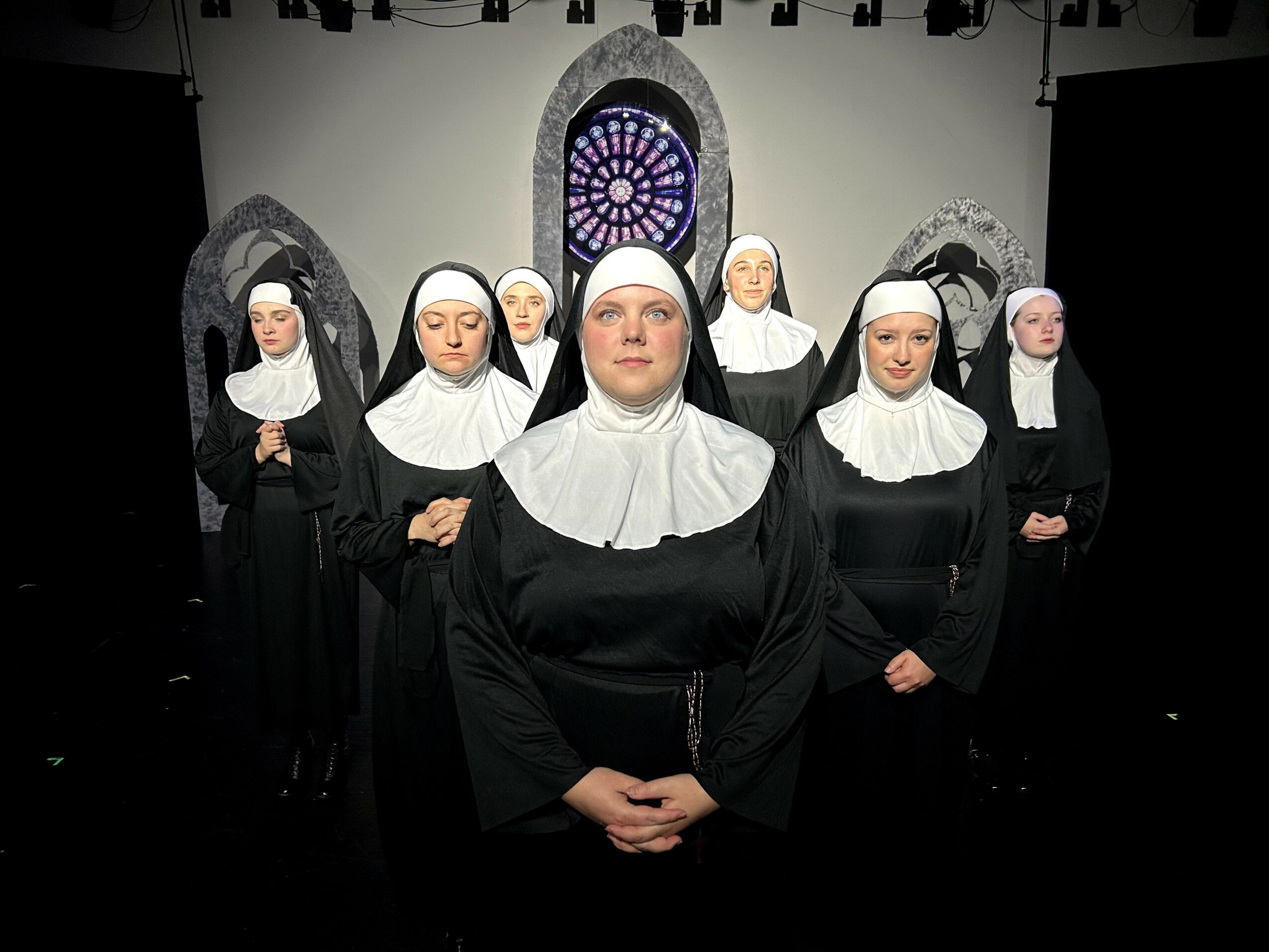Group of women dressed as nuns standing in front of a church window backdrop, facing forward with hands clasped.