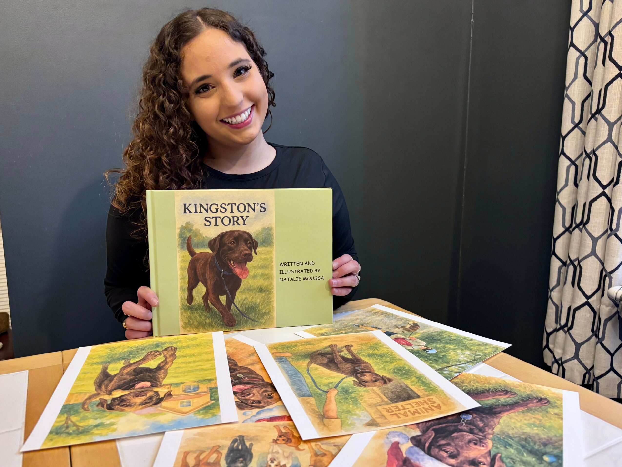Young woman with curly hair smiling and holding a book titled 'Kingston's Story' at a table with illustrated pages.