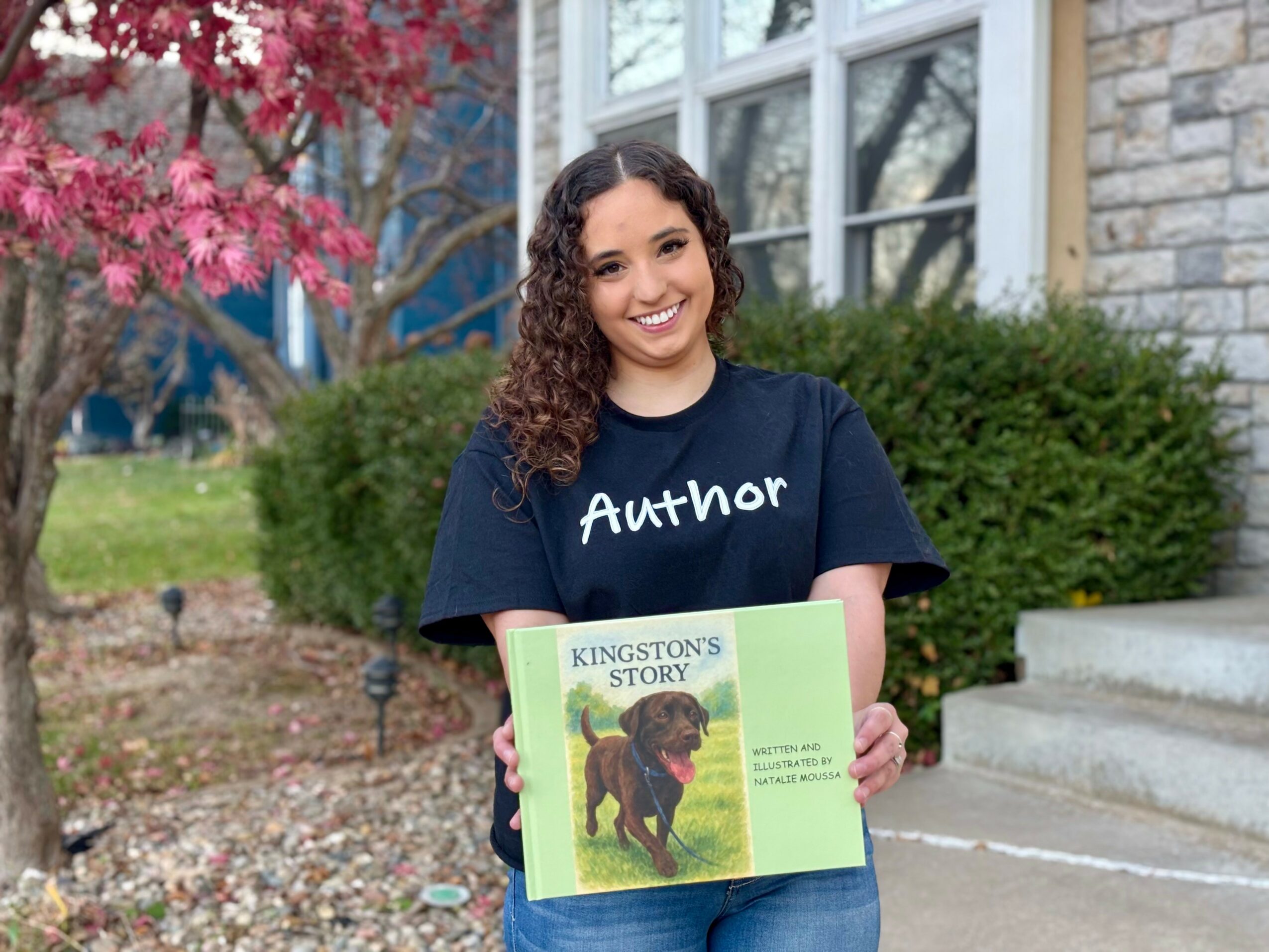 Smiling young woman holding a children's book outdoors, with a house, bushes, and pink flowering tree in background.