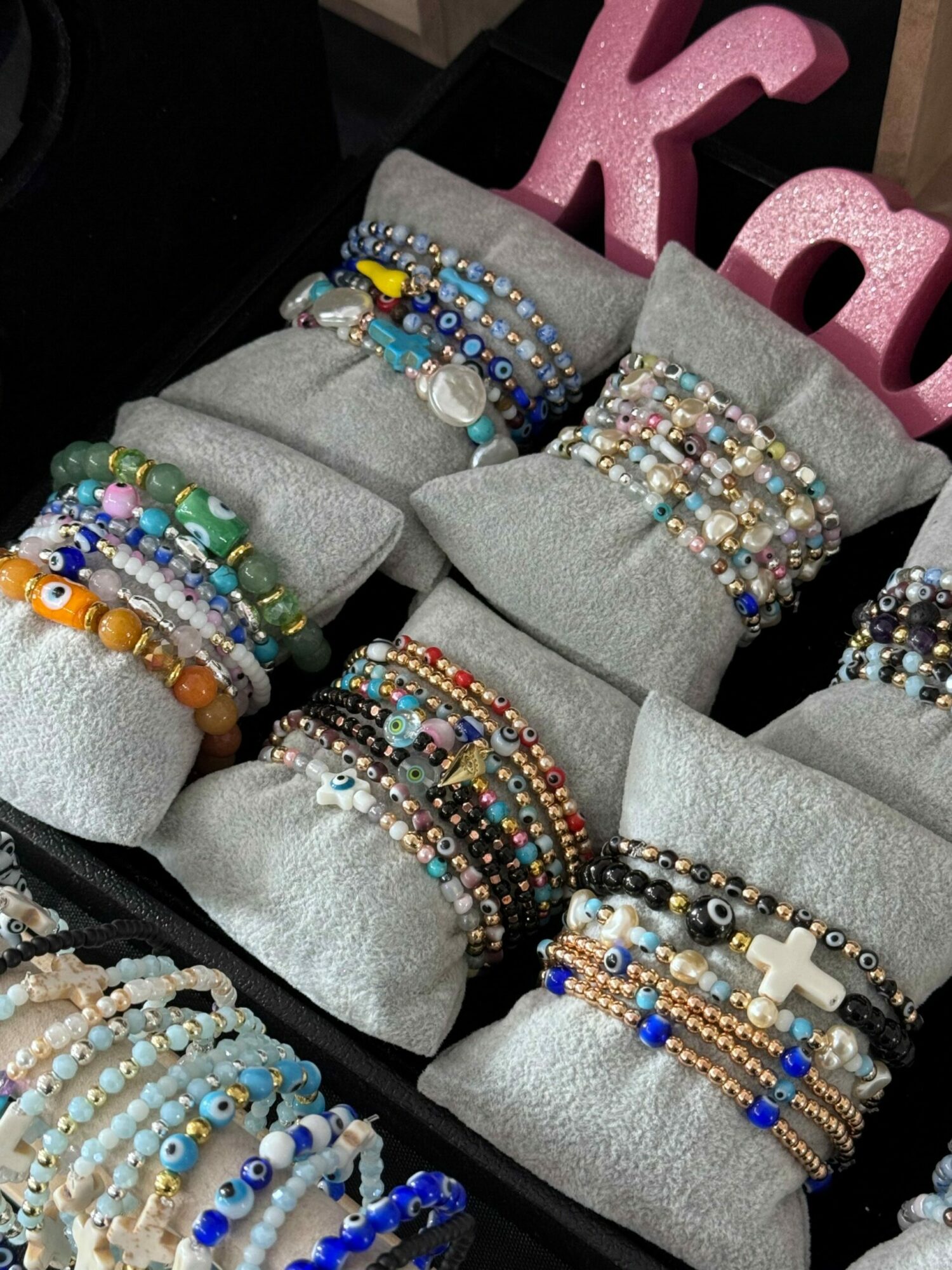 Multiple colorful beaded bracelets displayed on gray fabric rolls, with a pink decorative object in the background.