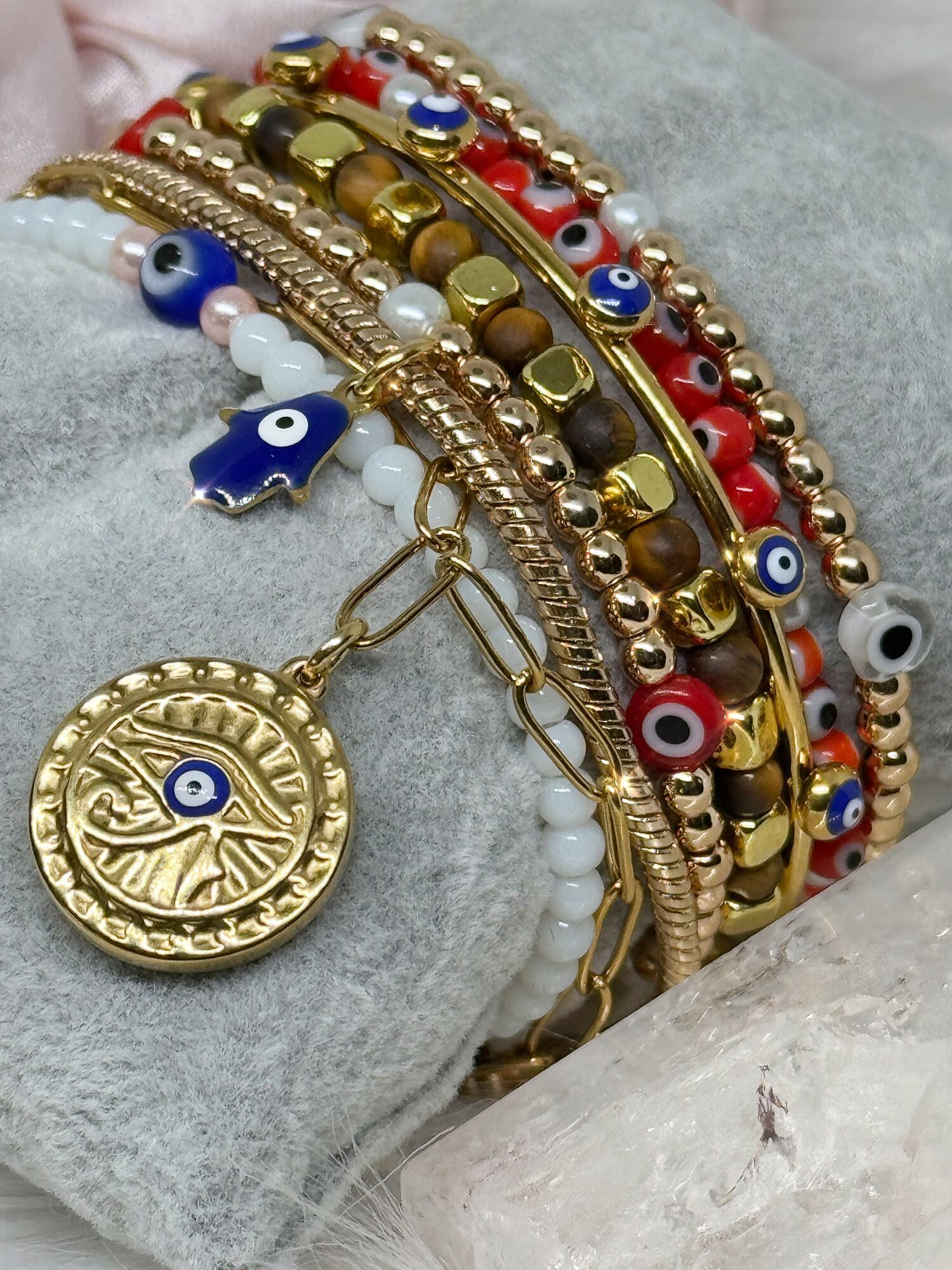 Multiple gold and red bracelets with eye-shaped charms and a round gold pendant with an eye symbol, resting on gray fabric.
