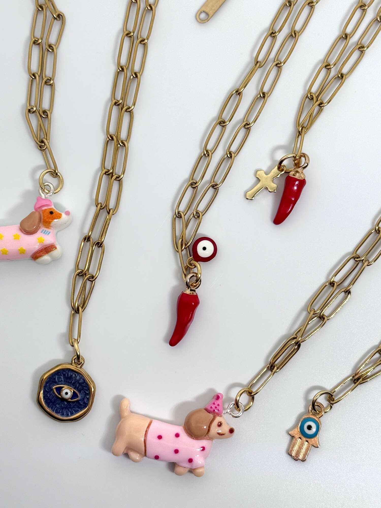 Collection of gold chains with colorful charms including animals, a chili pepper, a cross, and an eye, on a white background.