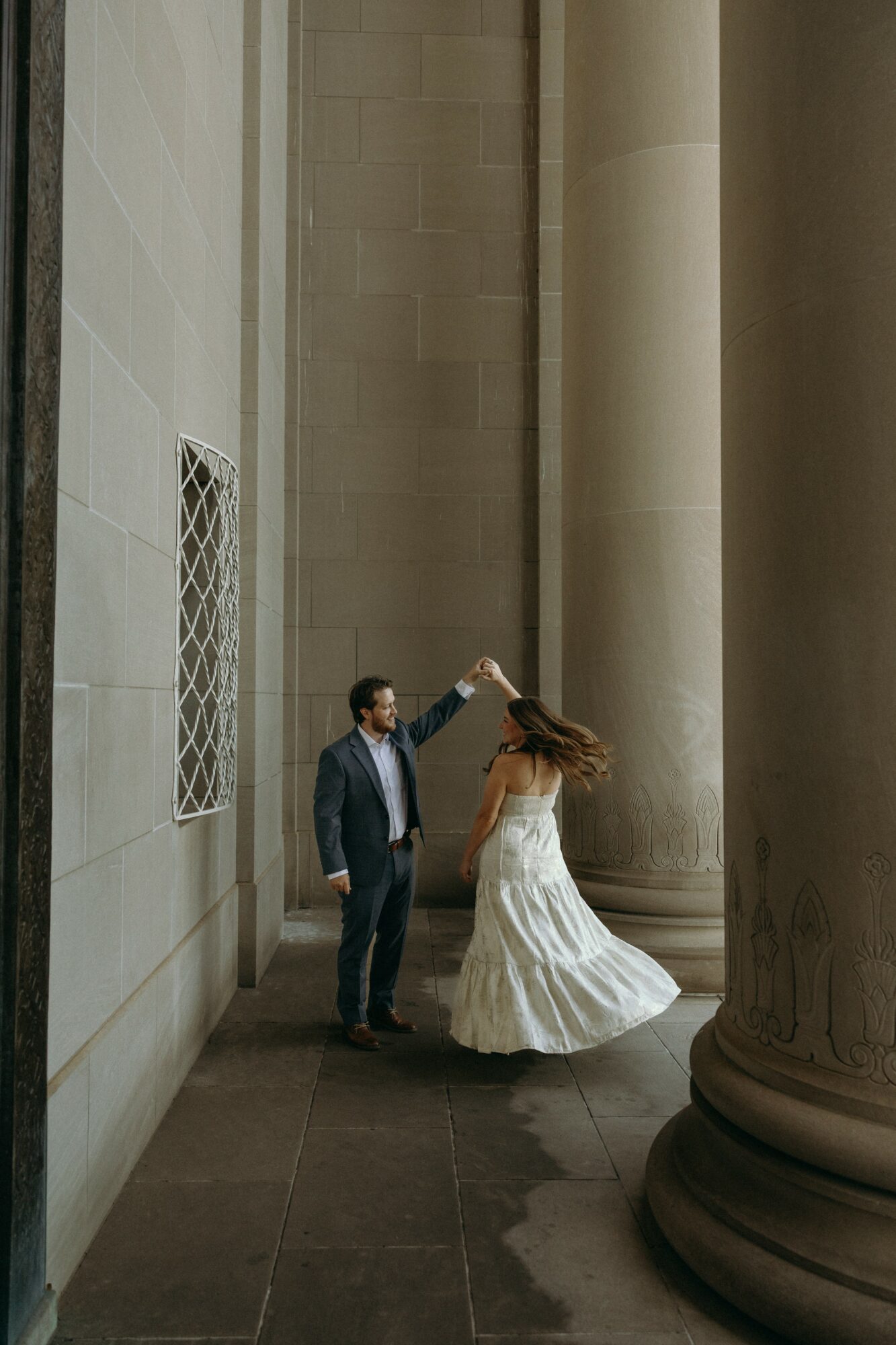 A couple dancing in a grand hall with tall columns and high ceilings, one person in a suit and the other in a flowing dress.