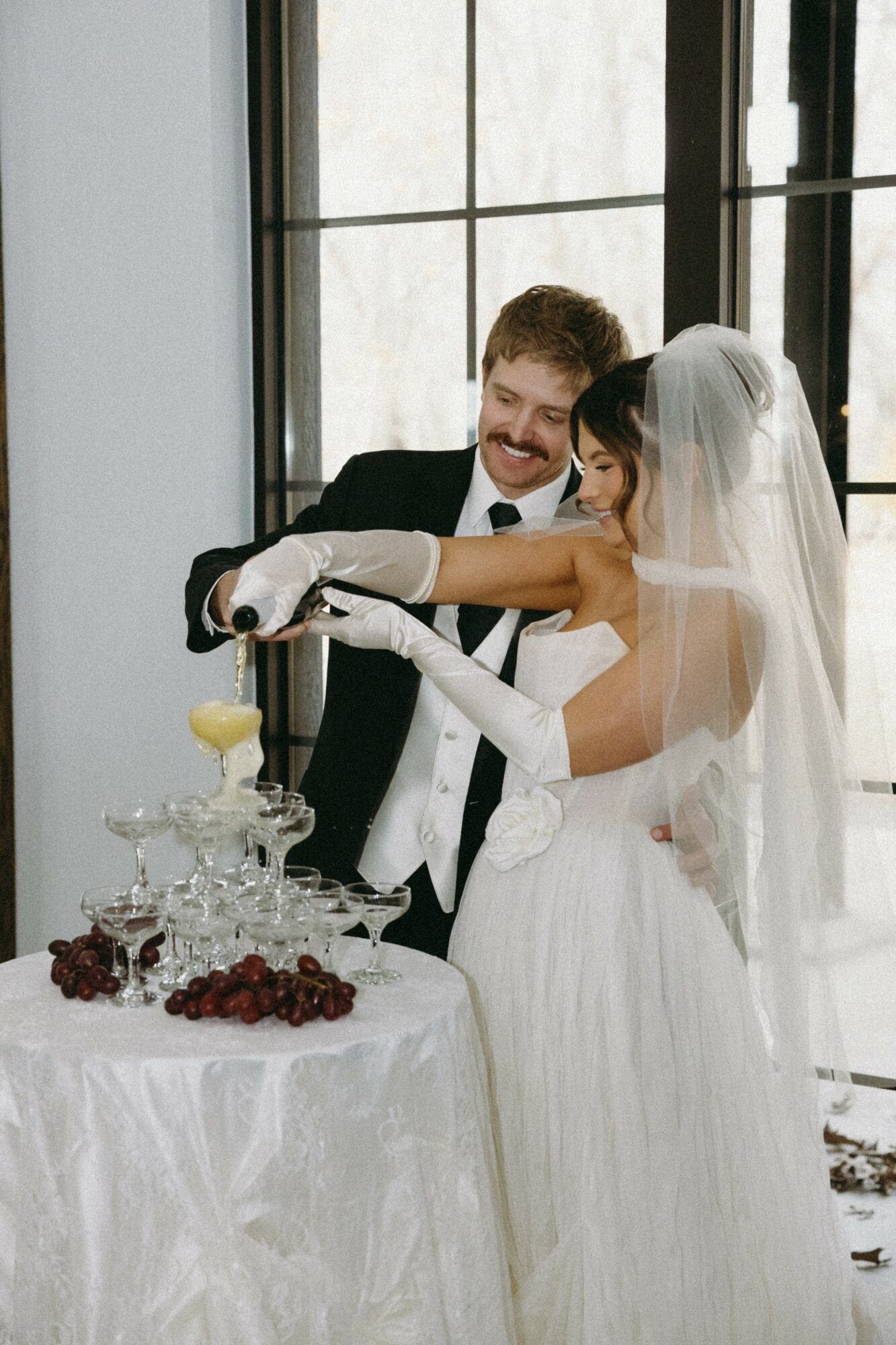 Bride and groom cut wedding cake together indoors near large windows, with a table of cake and fruit in front.