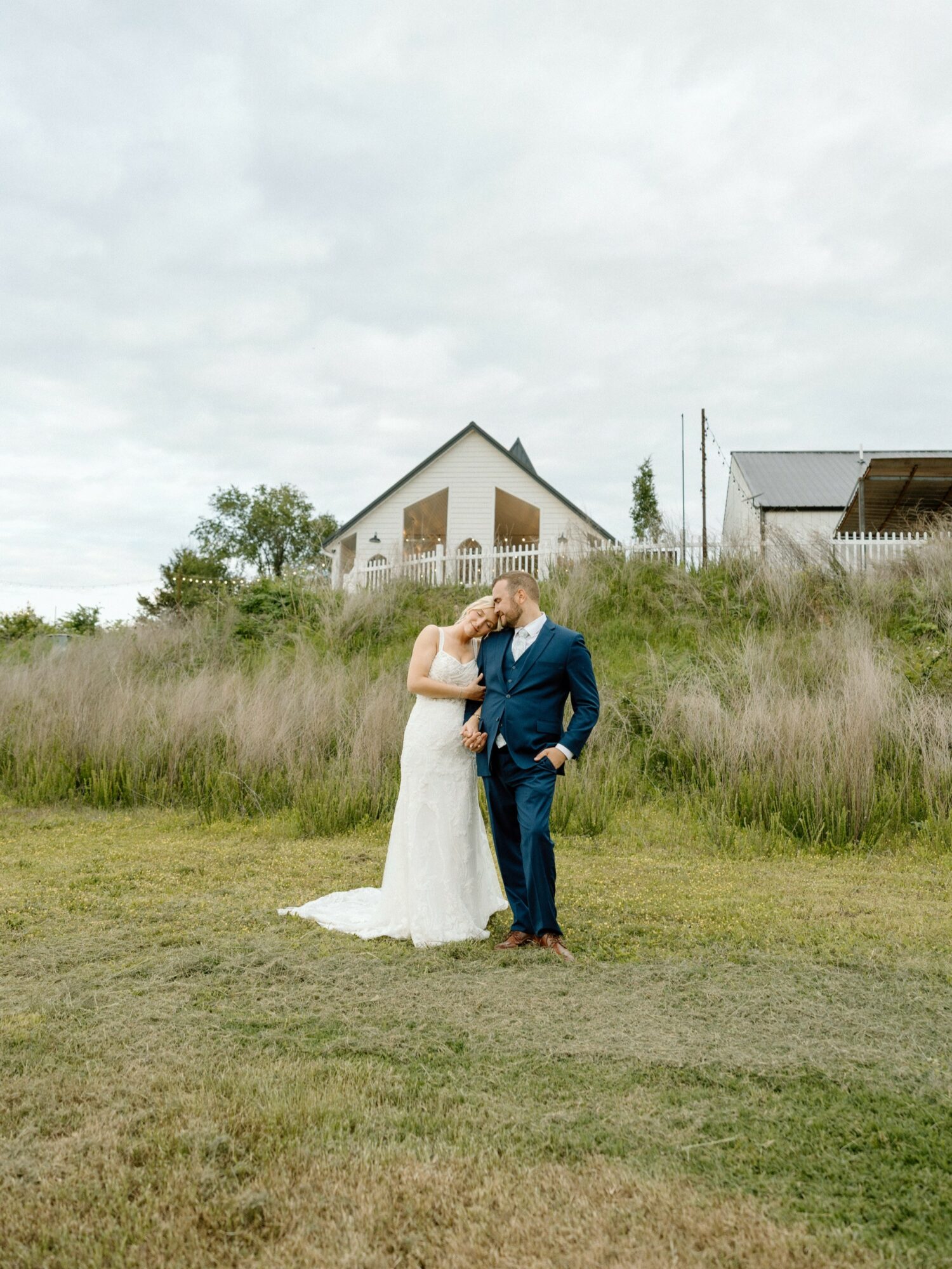 A bride and groom stand close together outdoors, with a house and grassy field in the background.