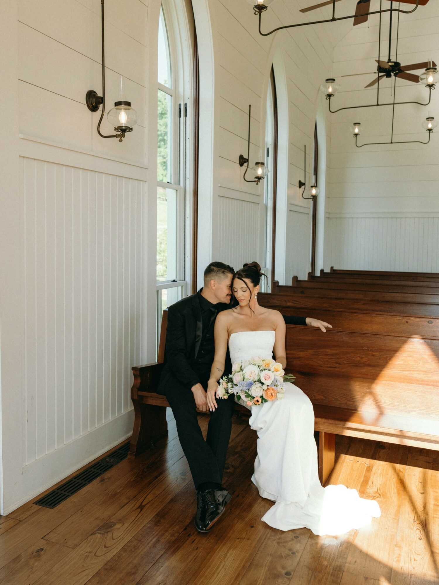Bride and groom sitting on a wooden bench inside a church, sharing a kiss, with sunlight streaming through windows.