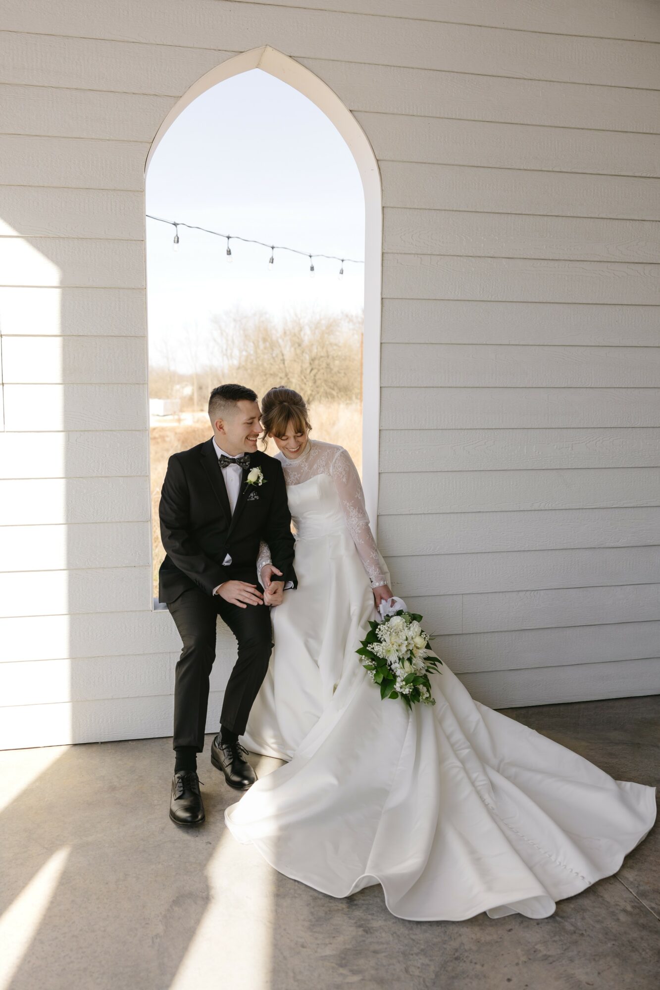 Bride and groom sit together in front of a white wall with an arched window, bride holding a bouquet, sunlight outside.