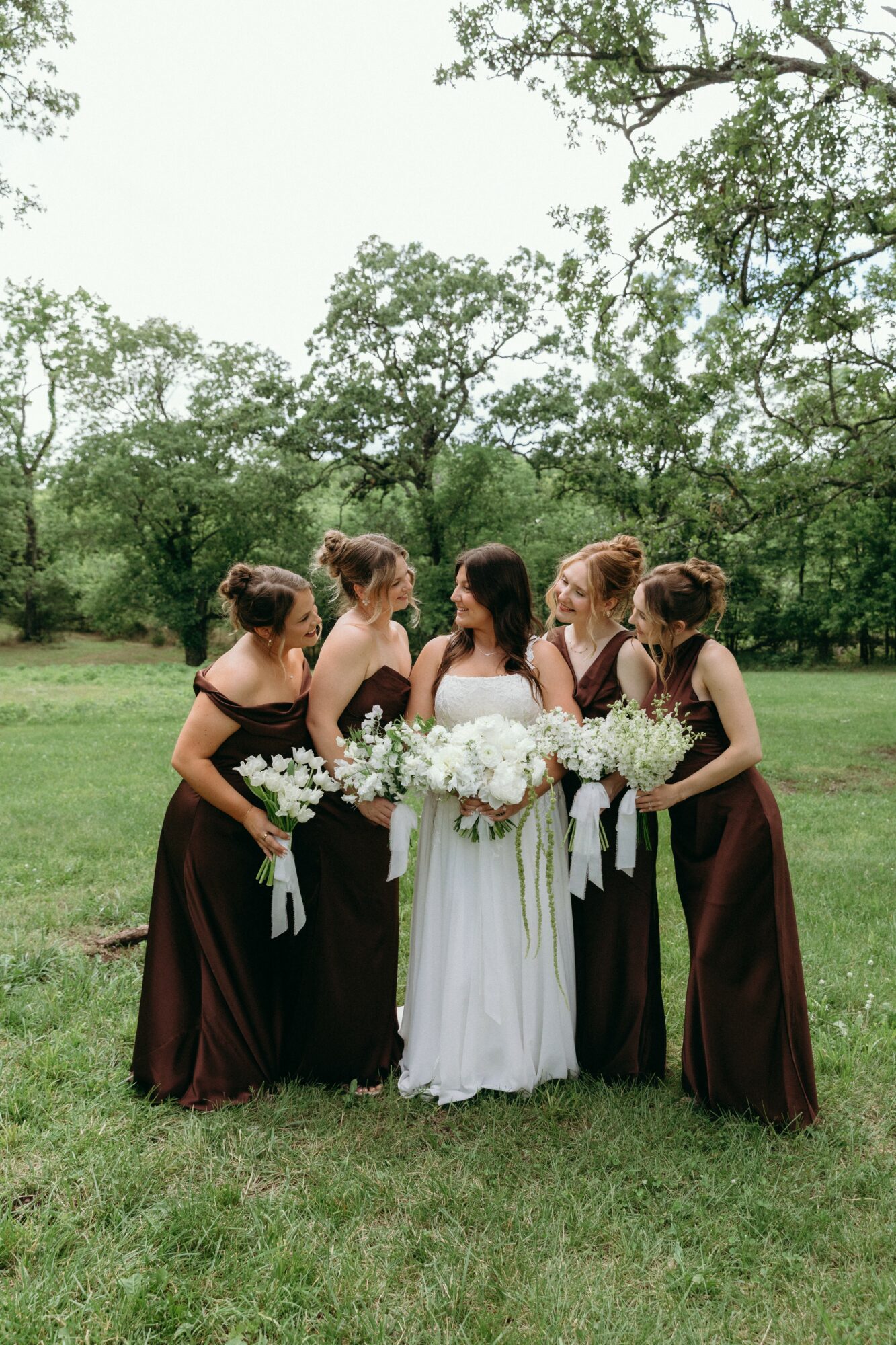 Five women in formal dresses standing outdoors on grass, holding bouquets, with trees in the background.