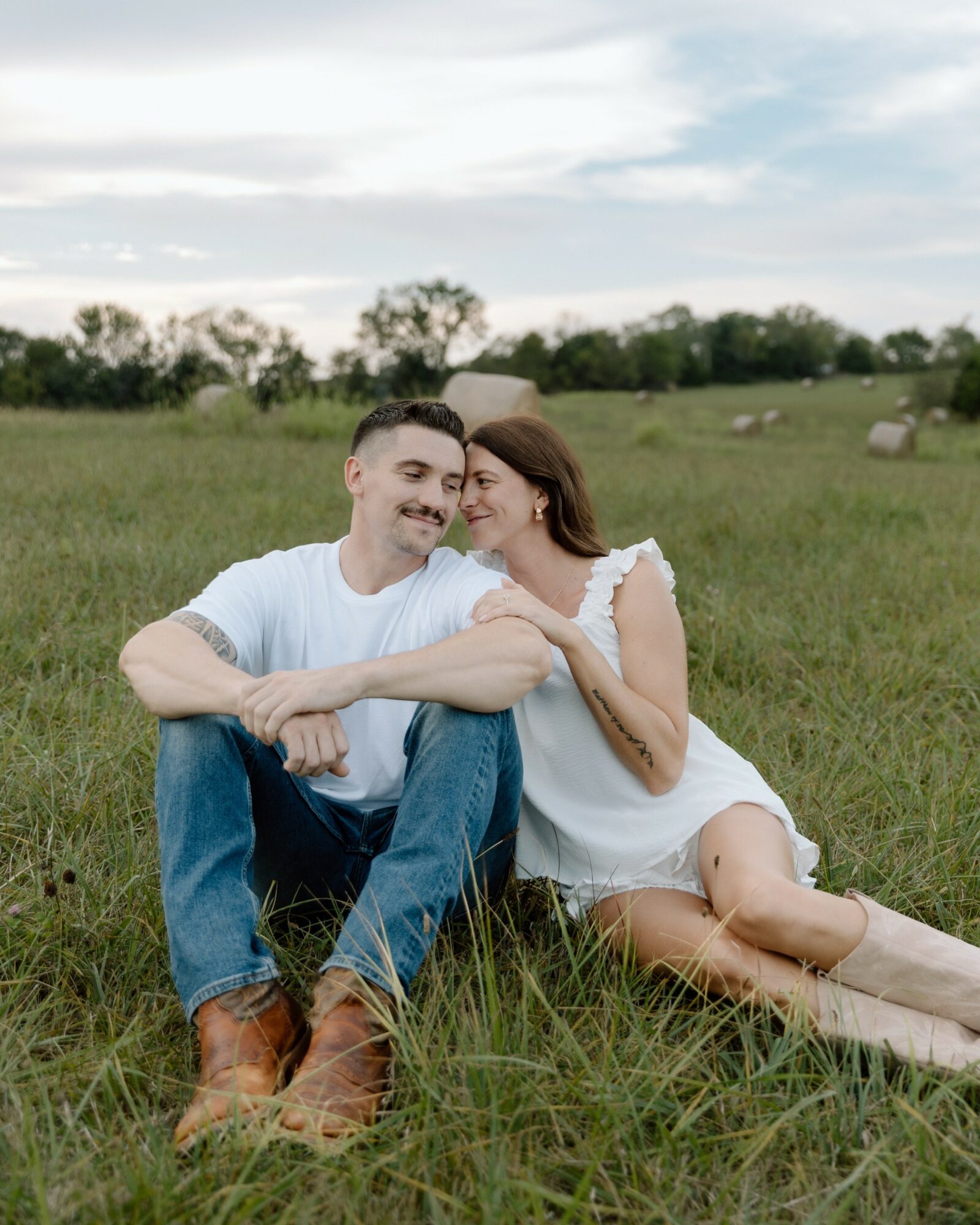 A smiling couple sitting on grass in a field, embracing each other, with trees and a cloudy sky in the background.