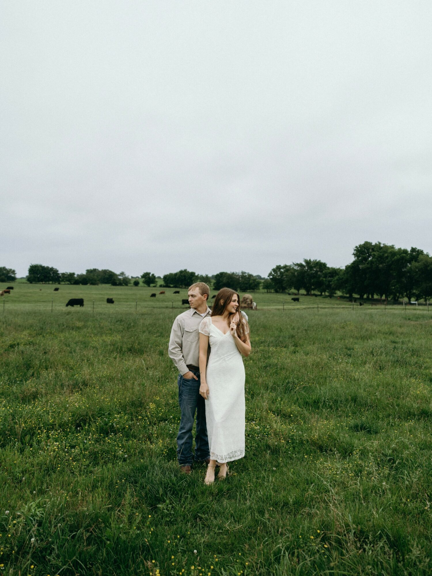 A couple standing in a green field with trees and grazing animals under a cloudy sky.