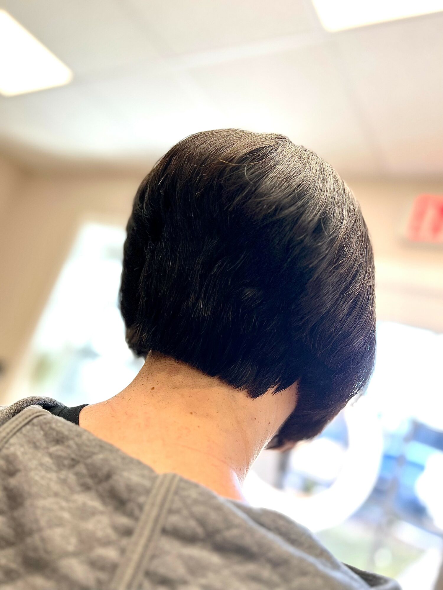 Back of person's head with short, dark hair in a layered bob style, indoors with bright lighting and windows.