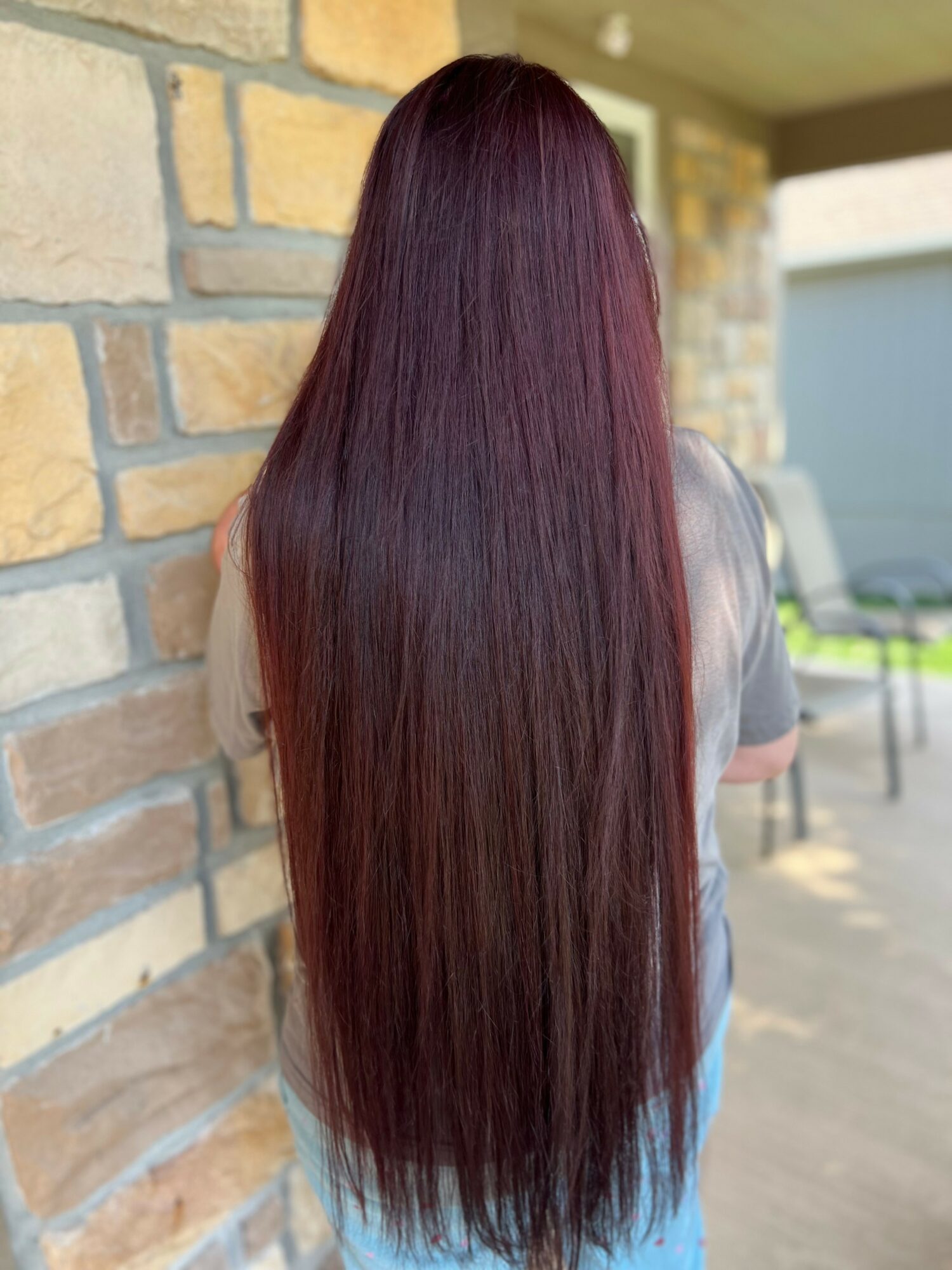 Person with long, straight, dark red hair standing outdoors near a stone wall, facing away from the camera.