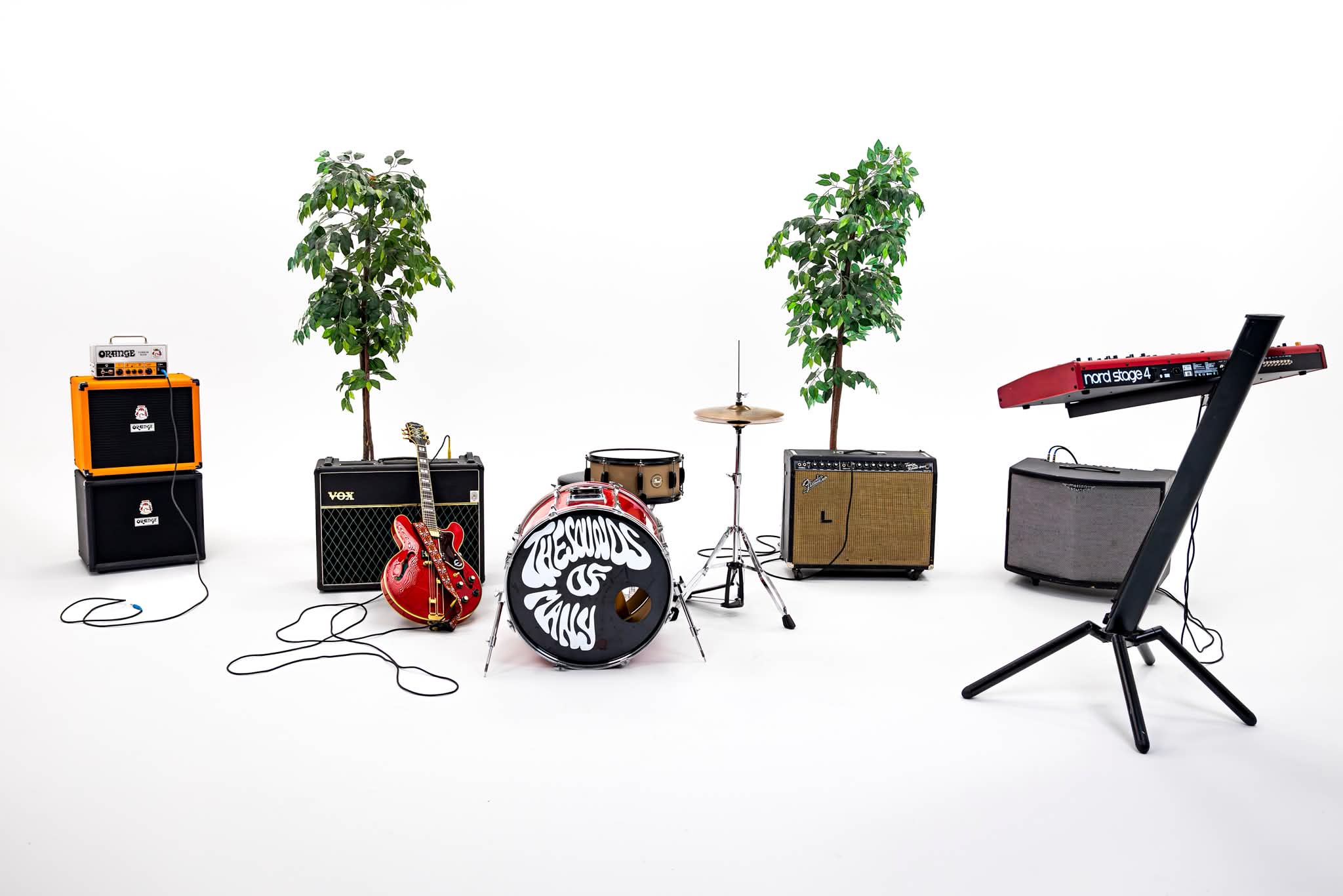 Music equipment including amplifiers, guitars, drums, keyboard, and a music stand arranged in a row on a white background.
