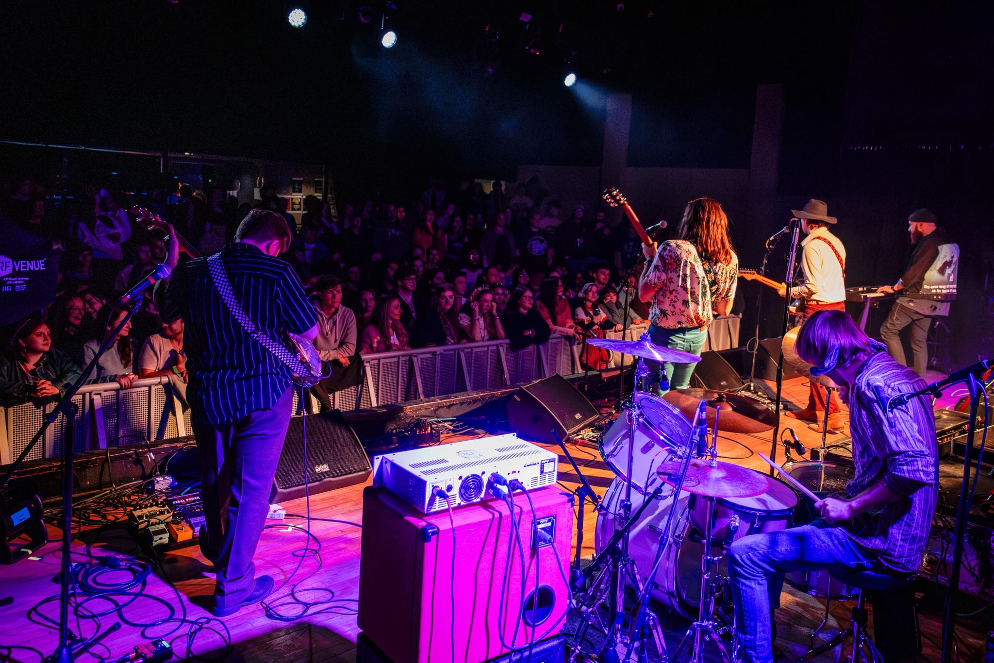 Band performing on stage with audience in front, stage lights shining, musical instruments visible, and a person playing drums.