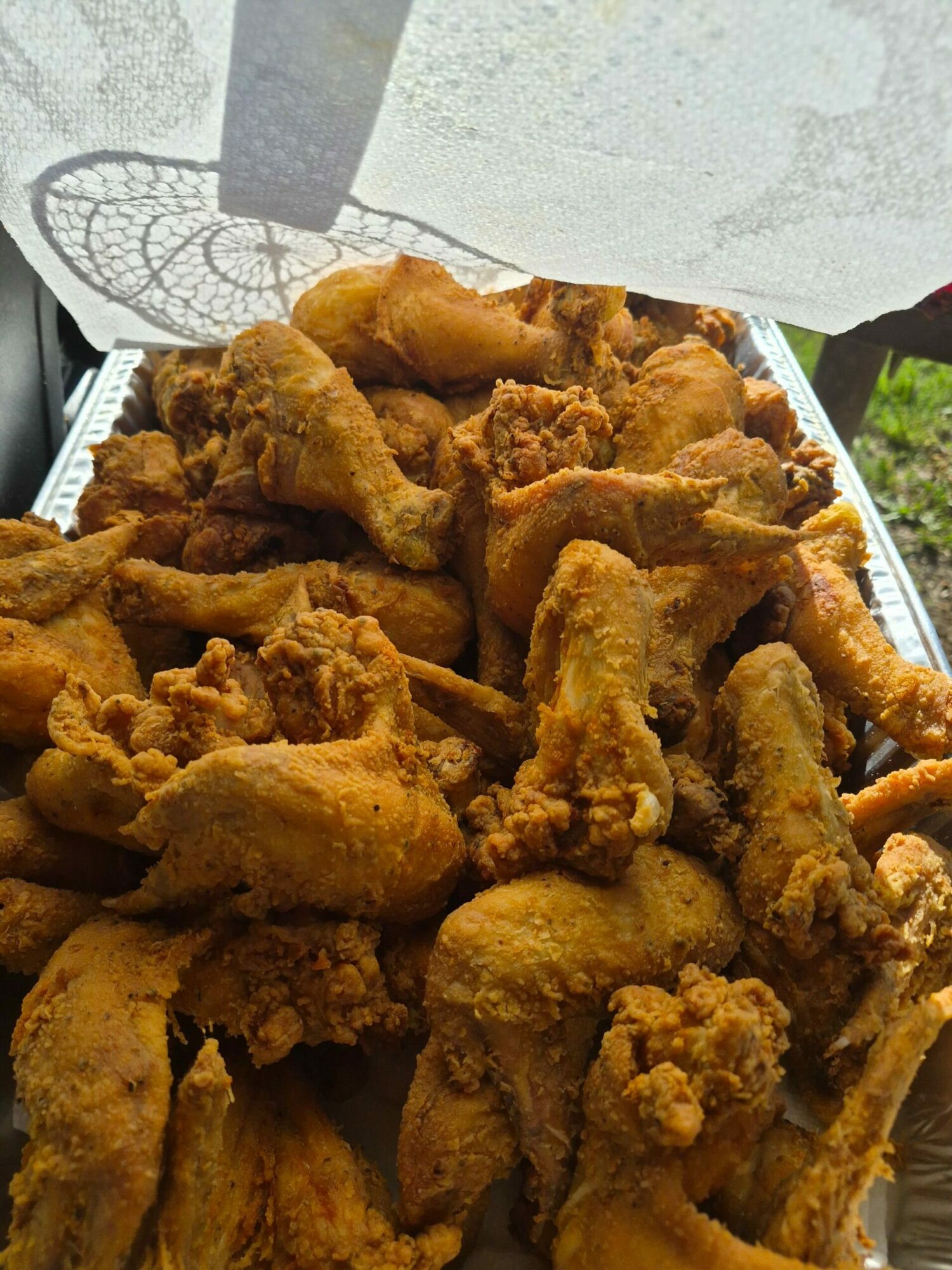 Pile of fried chicken pieces on a tray outdoors, with a paper towel and greenery in the background.