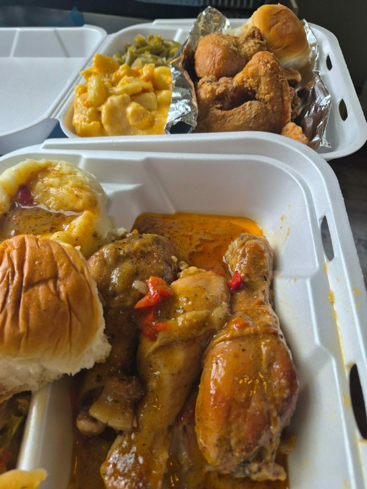 Two takeout containers with fried chicken, macaroni and cheese, and a chicken drumstick in sauce, viewed from above.