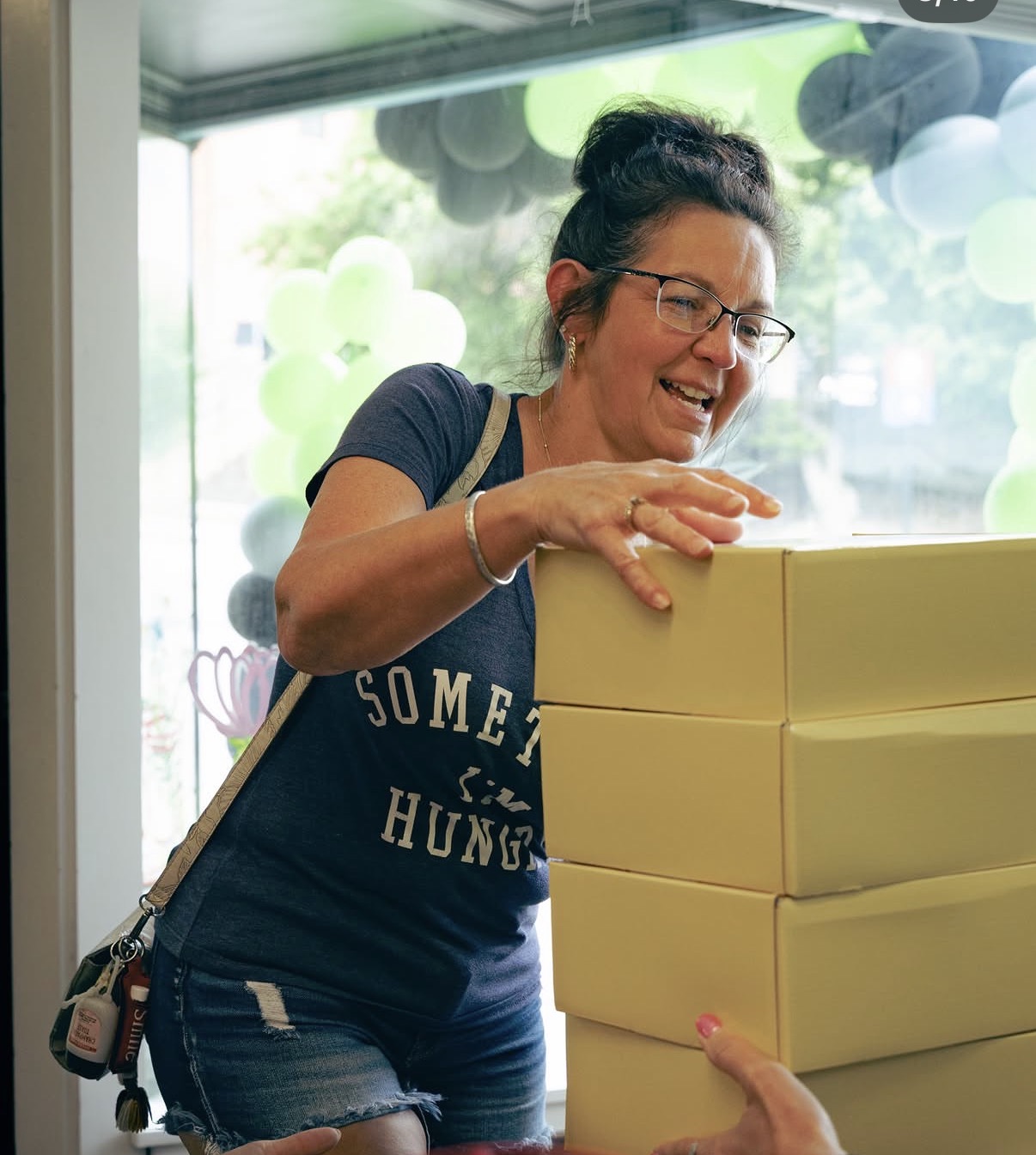 Woman with glasses smiling, leaning on a stack of yellow boxes, wearing a navy T-shirt and denim shorts, indoors with balloons outside