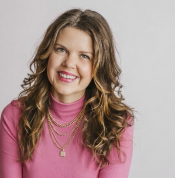 Woman with wavy hair smiling, wearing a pink turtleneck and layered necklaces, against a plain background.