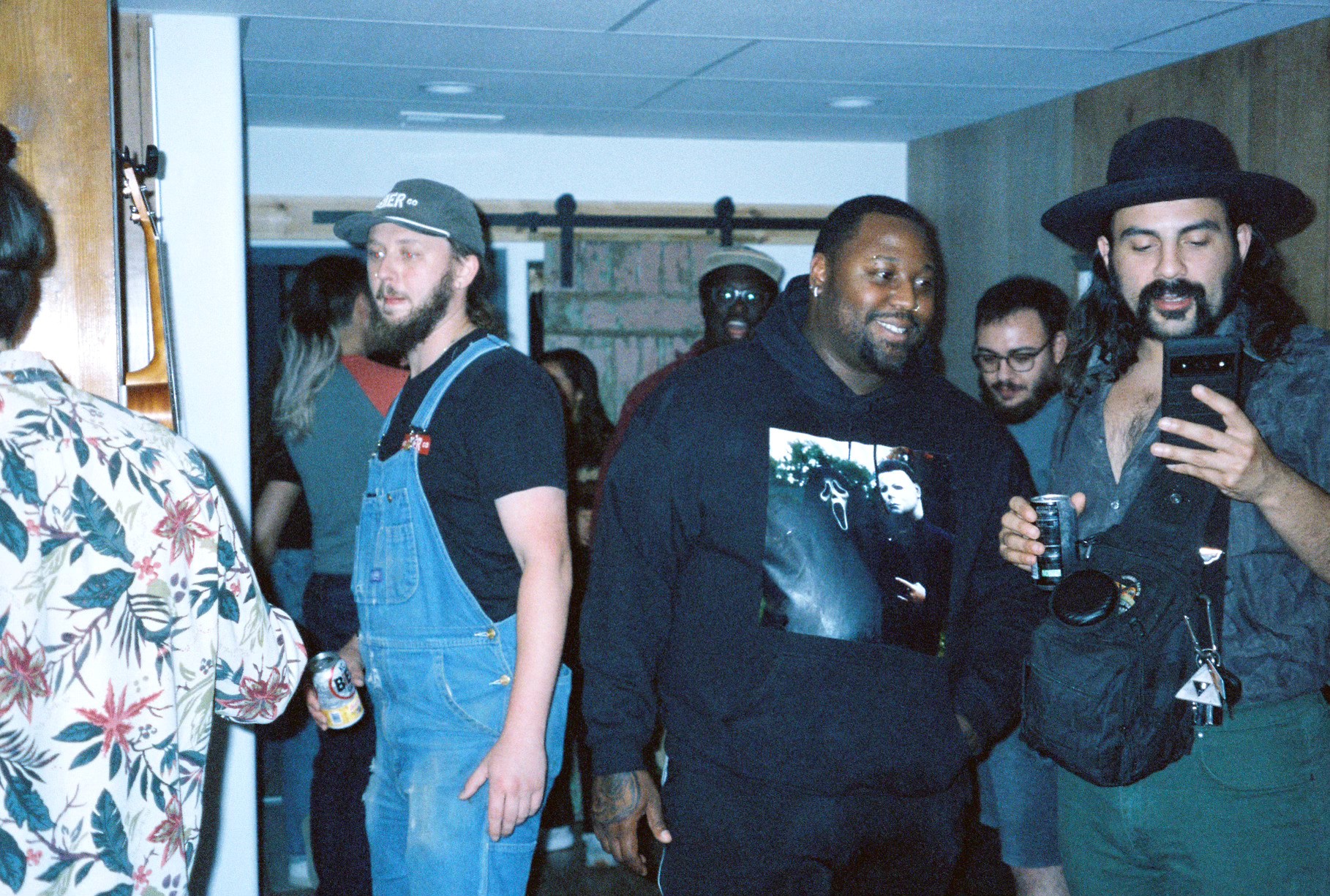 Group of people in an indoor space, some looking at a phone, others talking or observing, with a person in floral shirt on the left.