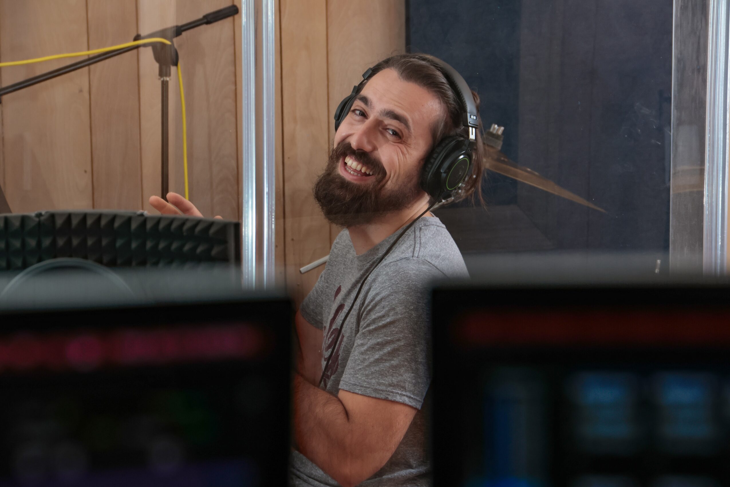 Man with headphones smiling in a recording studio, seen through a glass window, with audio equipment in foreground.