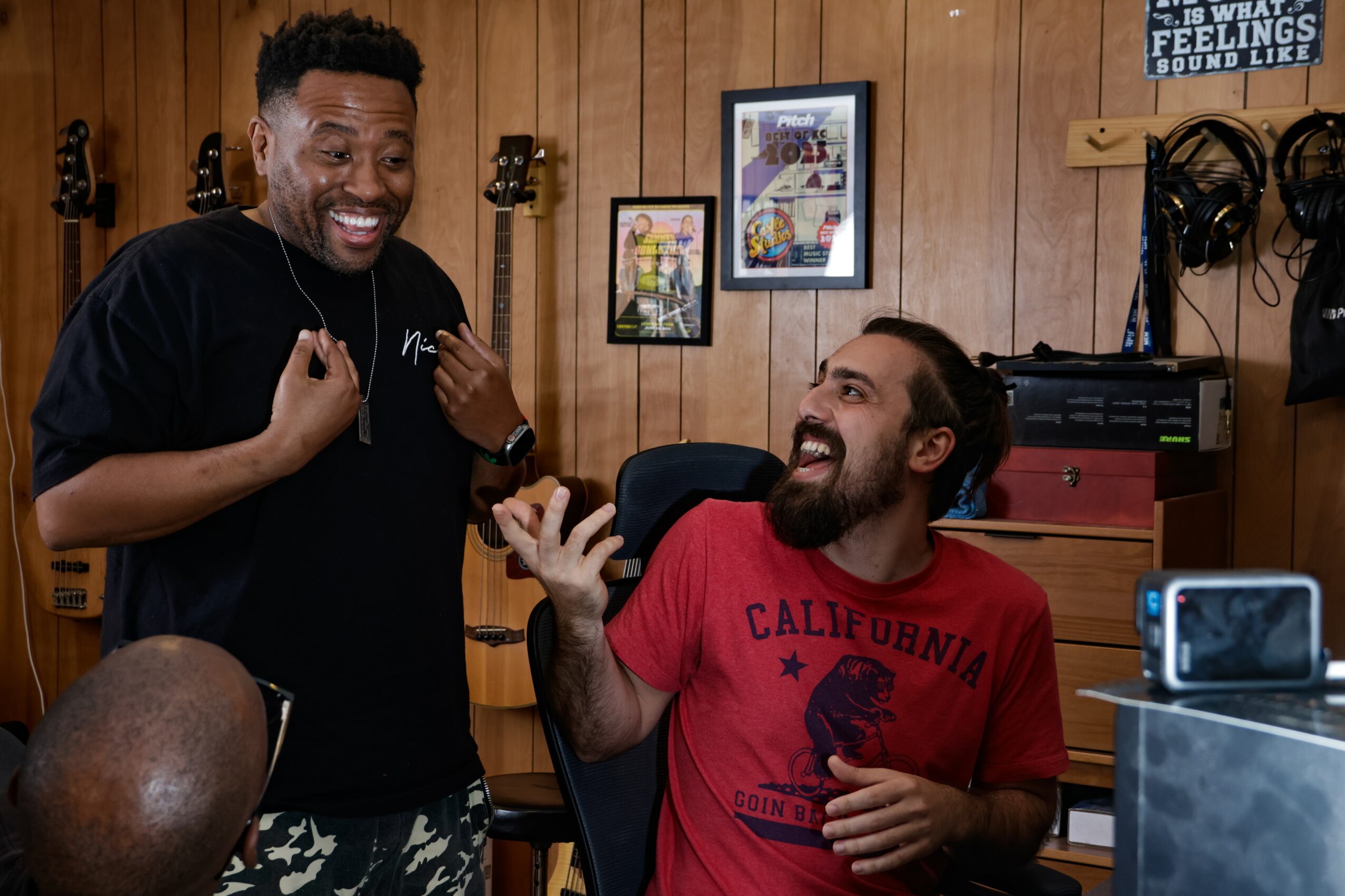 Two men are smiling and talking in a music studio with wooden walls, guitars, and framed pictures behind them.