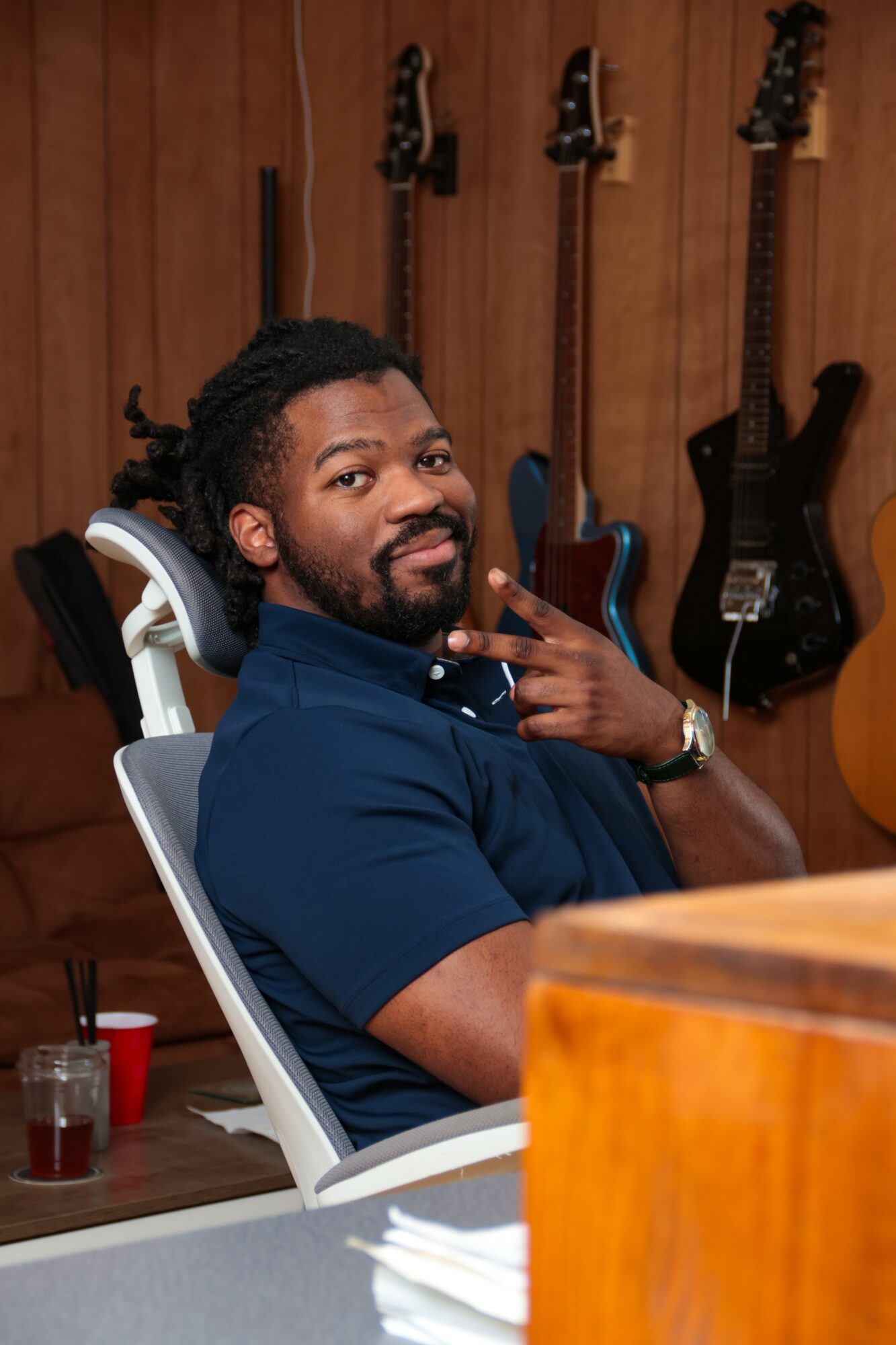 Man with dreadlocks and beard sitting in a chair, making a peace sign, with guitars hanging on a wooden wall behind him.