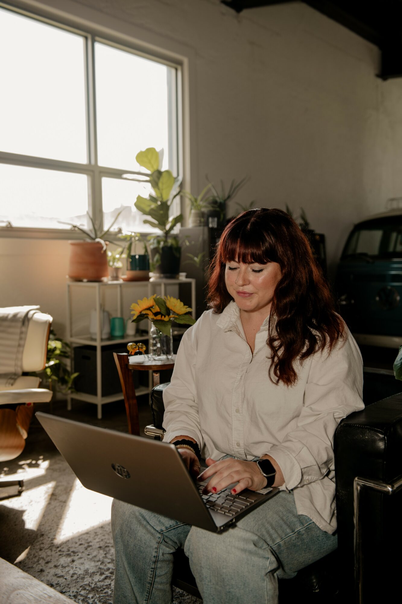 Woman with long dark hair and bangs working on a laptop indoors, sunlight streaming through a window, plants on shelves behind her.