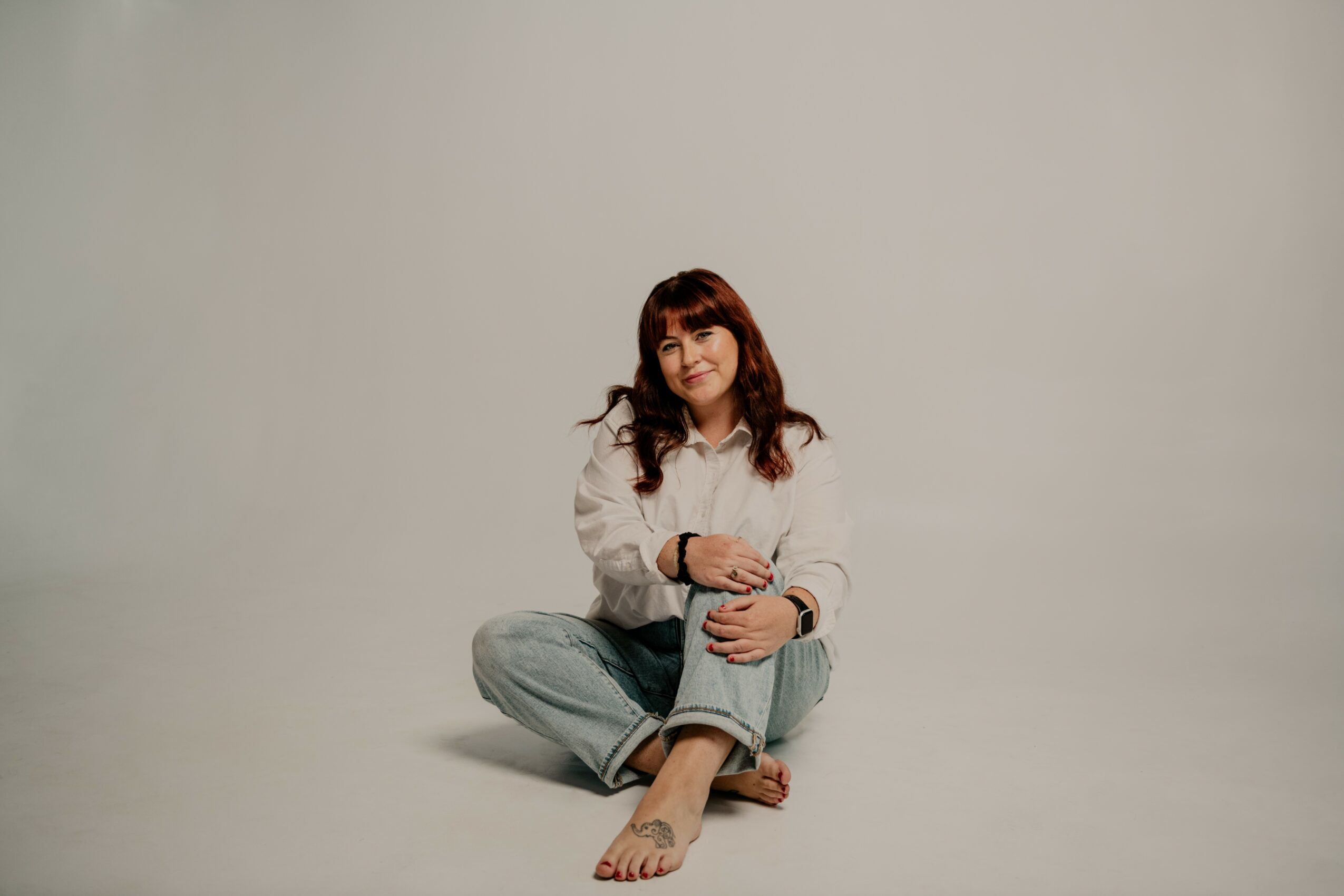 Woman sitting cross-legged on the floor, smiling, wearing a white shirt and jeans, against a plain background.