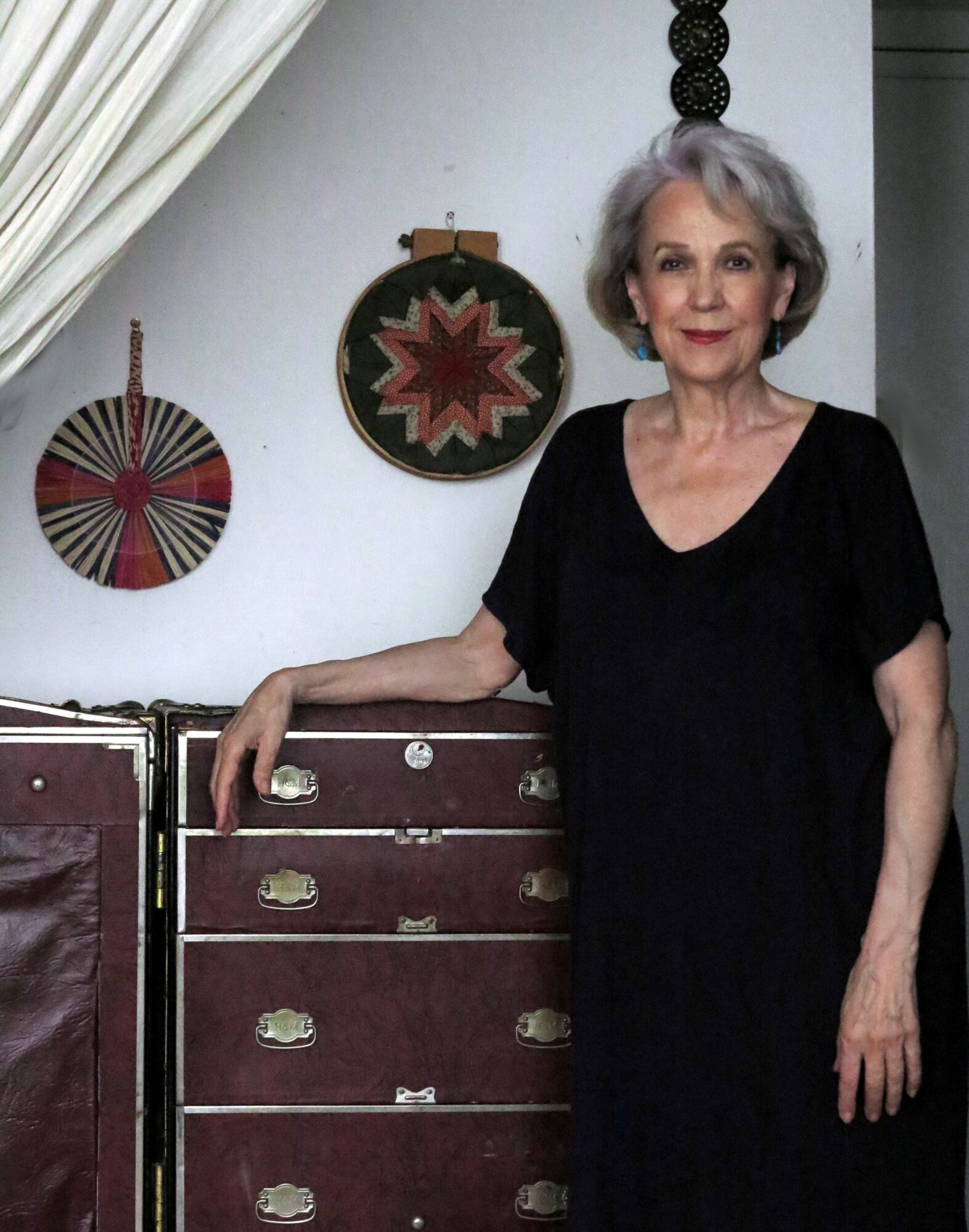Older woman with gray hair in a black dress standing indoors, with decorative wall hangings behind her.