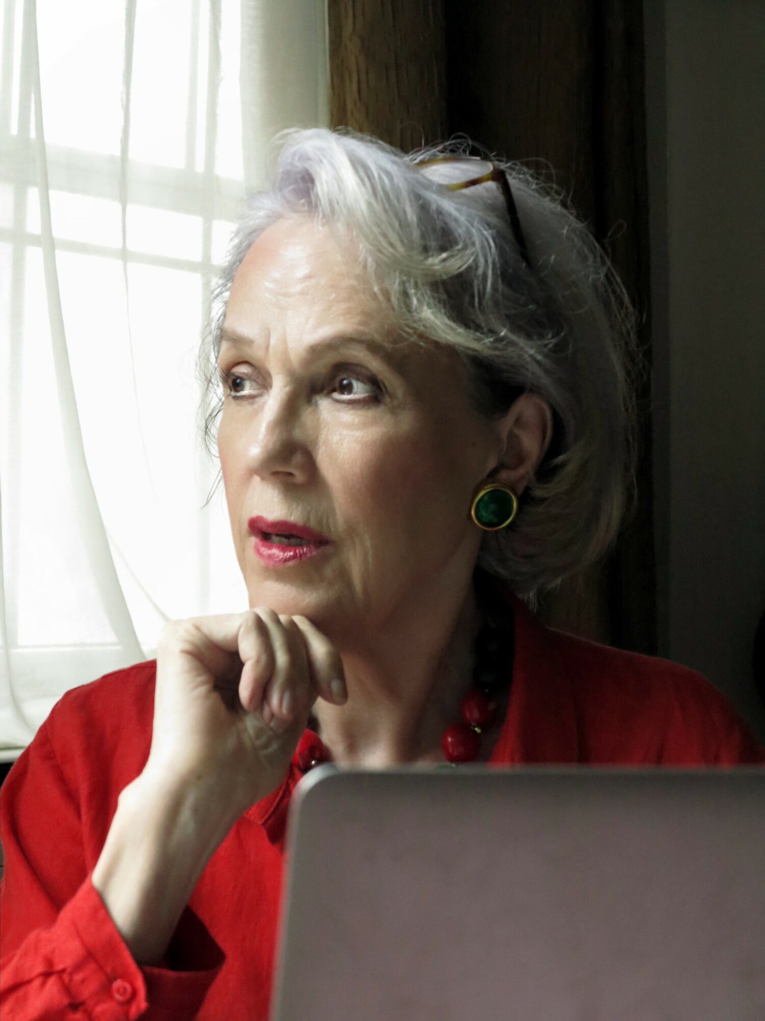 Elderly woman with gray hair, wearing red, sitting near window, looking thoughtfully outside, hand near chin, laptop in foreground.