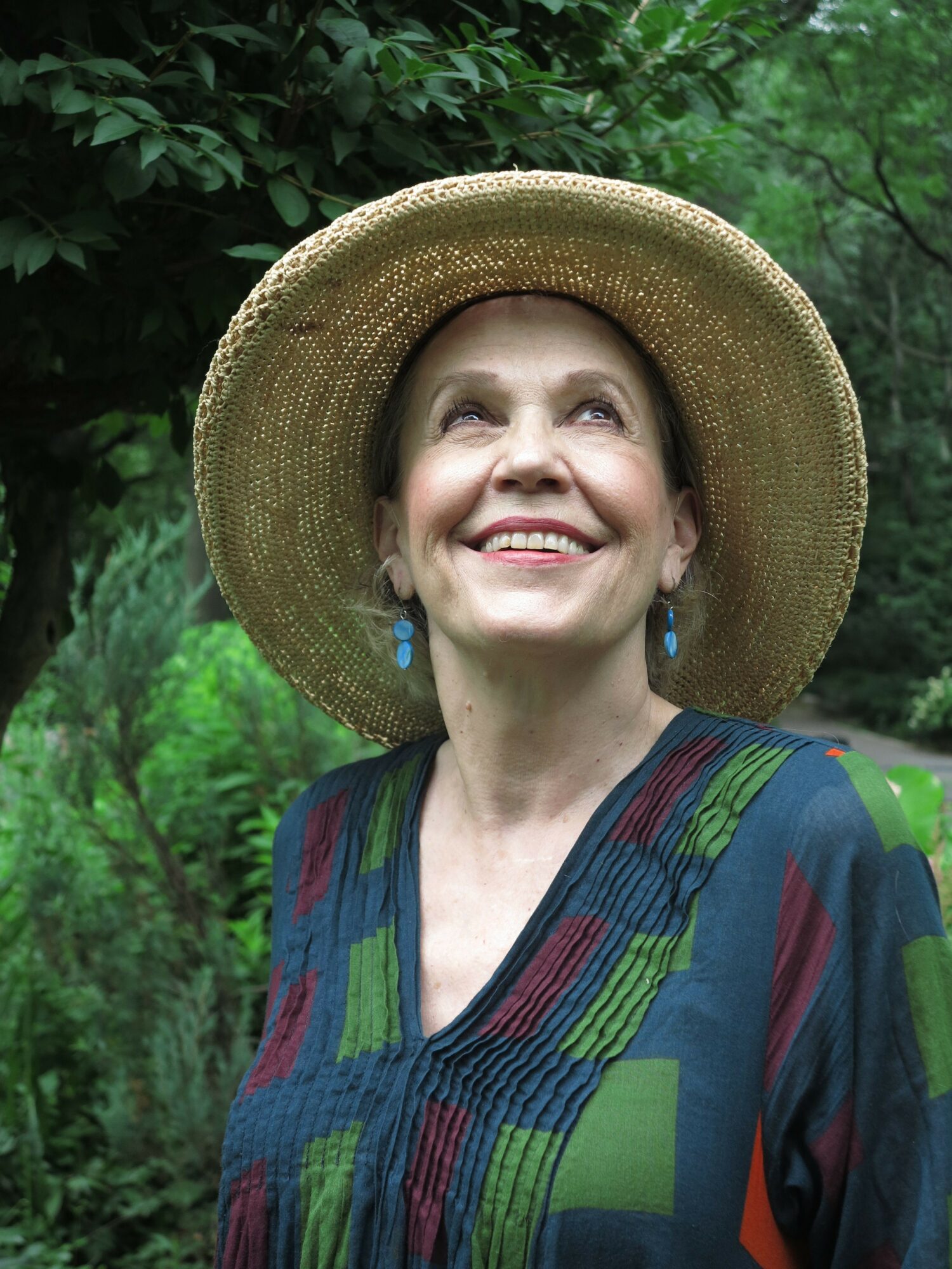 Woman wearing a large straw hat and colorful patterned top, smiling outdoors with green foliage background.