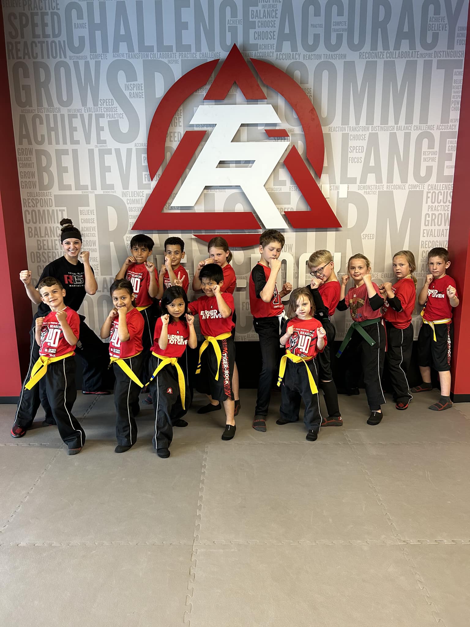 Group of children in martial arts uniforms and red shirts posing in front of a wall with a large logo and motivational words.
