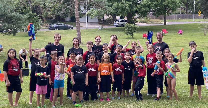 Group of children and adults outdoors on grass, some holding colorful objects, with trees and cars in background.