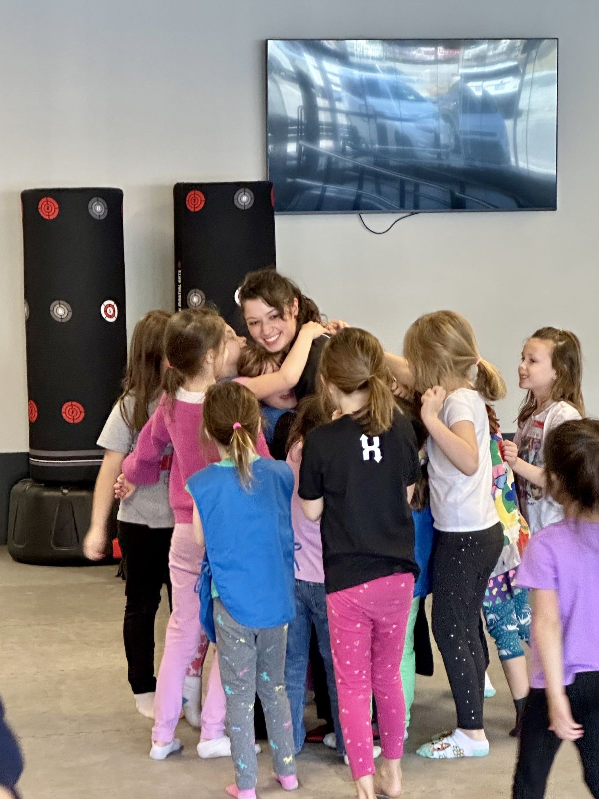 Group of children gathered around a smiling woman in a room with a large TV and speakers, engaging in an activity.