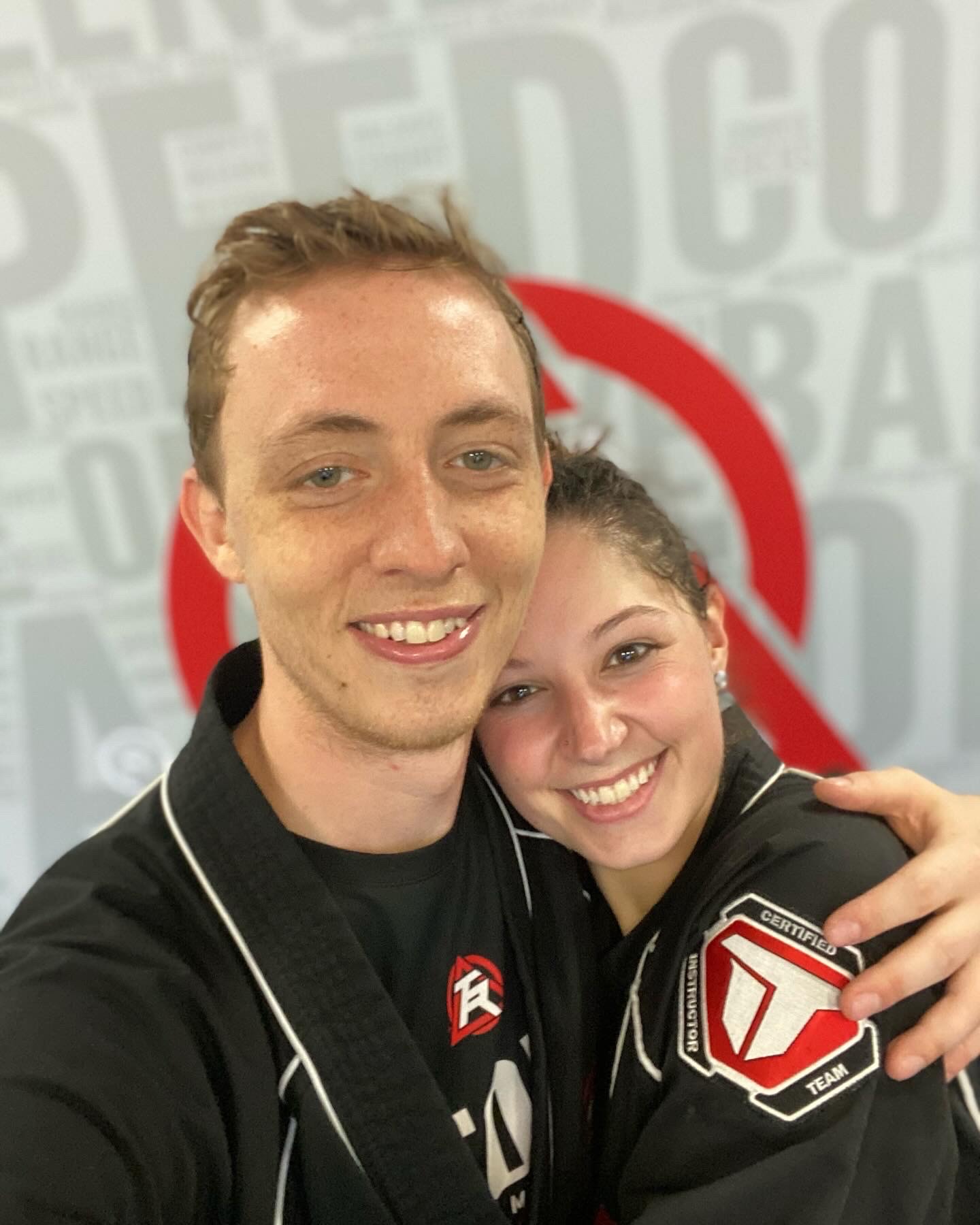 Two smiling people hugging, one wearing a black jacket with patches, in front of a background with text and a red circle.