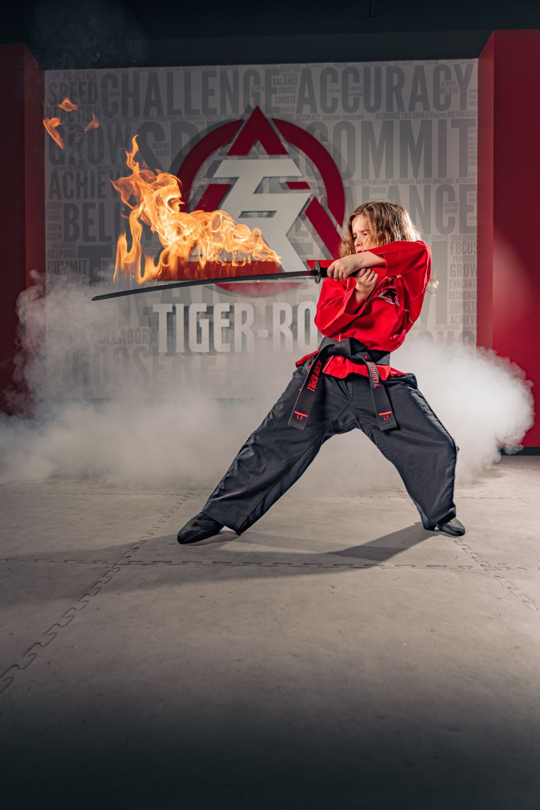 Child in martial arts uniform performs a fire-breathing stunt with flames and smoke, in front of a backdrop with words and a logo.