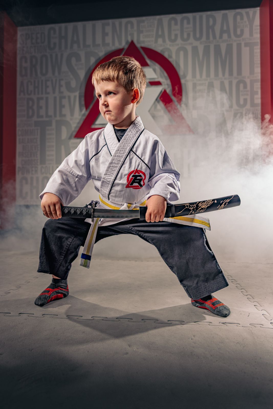 Young boy in martial arts uniform holding a sword, sitting in a martial arts pose with a smoky background and motivational words.