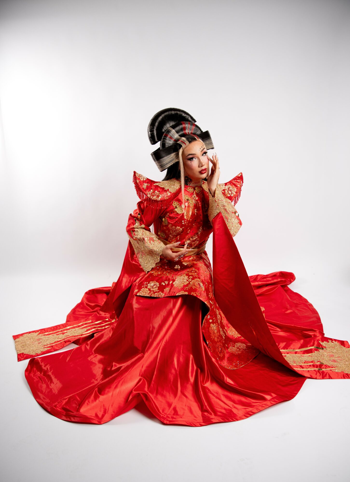 Woman in traditional red and gold dress with elaborate headpiece, sitting on the floor against a plain background.