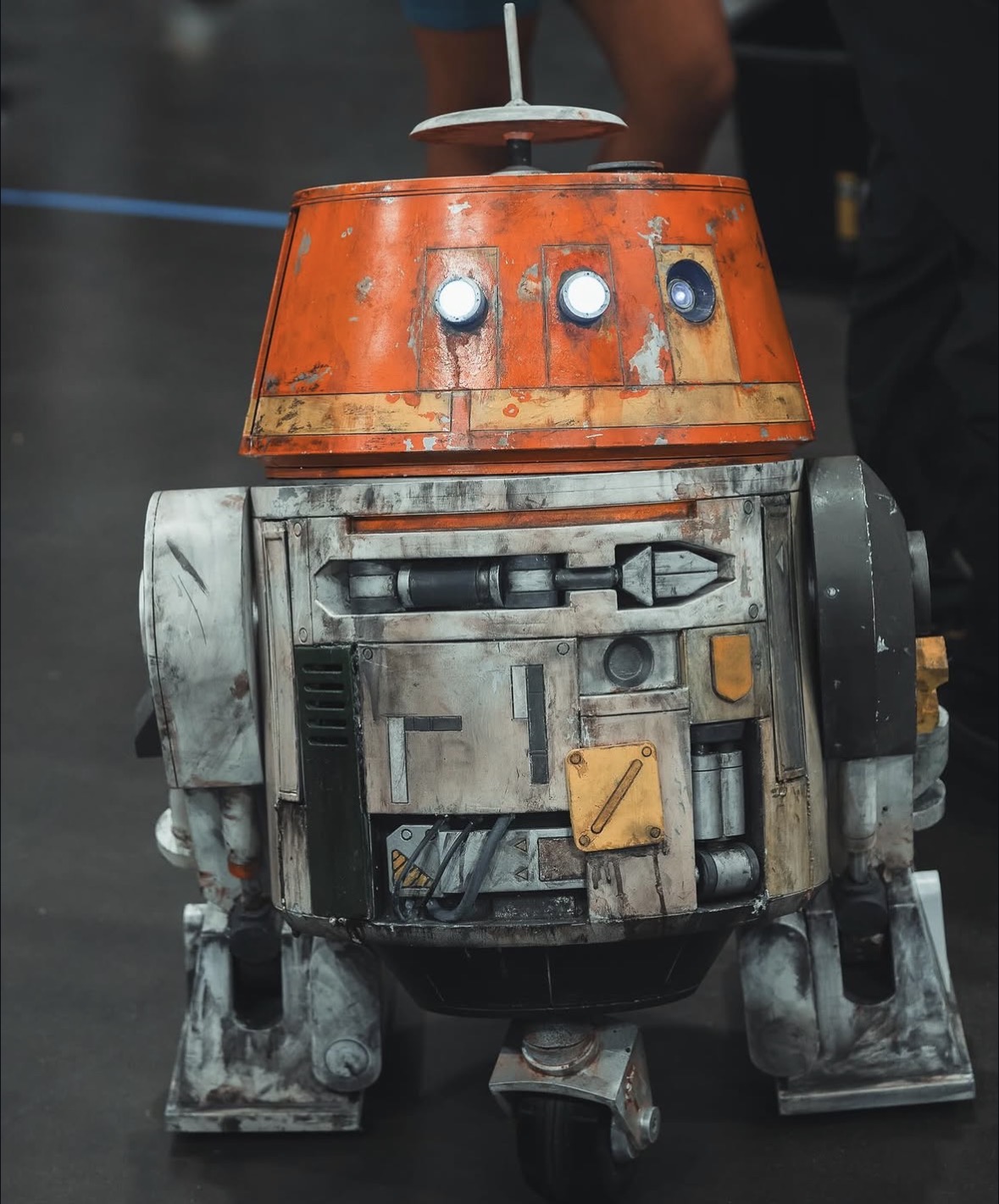 A robot with an orange top and gray body, standing on a dark floor, with people in the background.