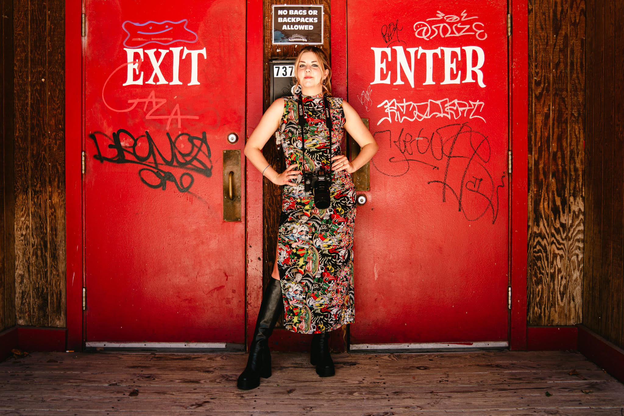 Woman standing between red exit and enter doors with graffiti, hands on hips, wearing a floral dress, holding a camera.