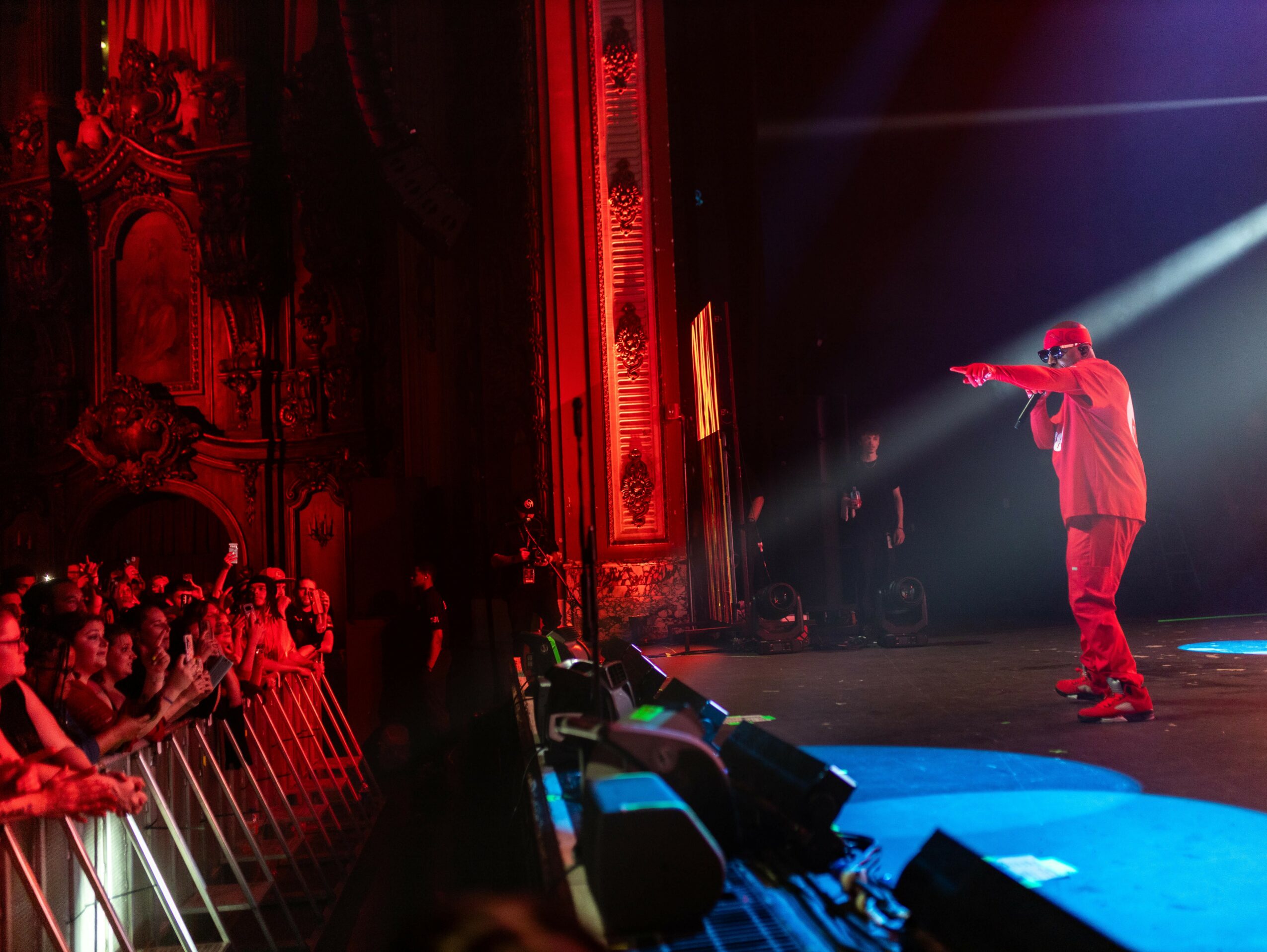 Performer on stage with microphone, audience watching, ornate theater interior, red lighting, stage lights shining, people in front row.