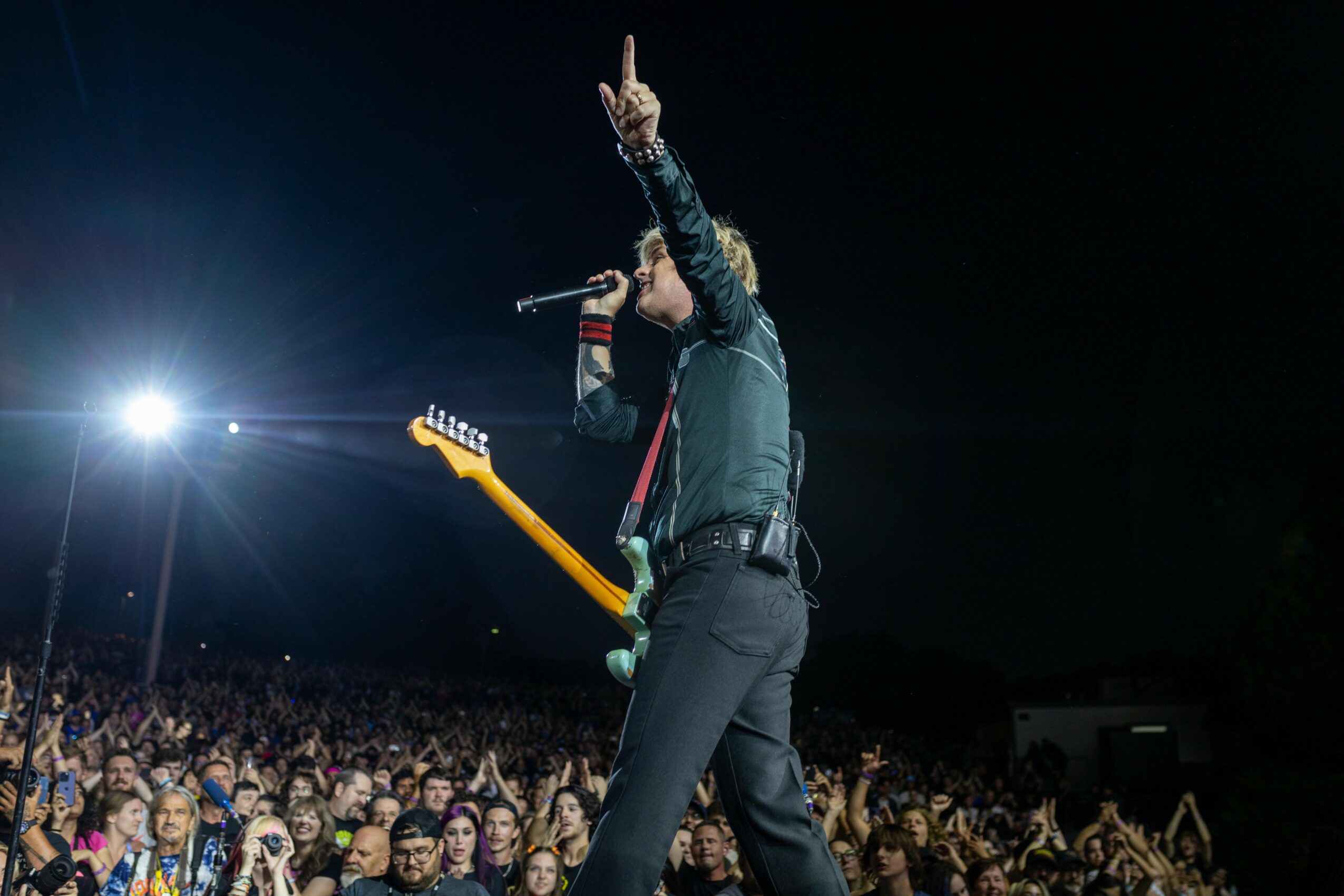 Performer on stage with a guitar, holding a microphone, raising one hand, in front of a large audience at night.