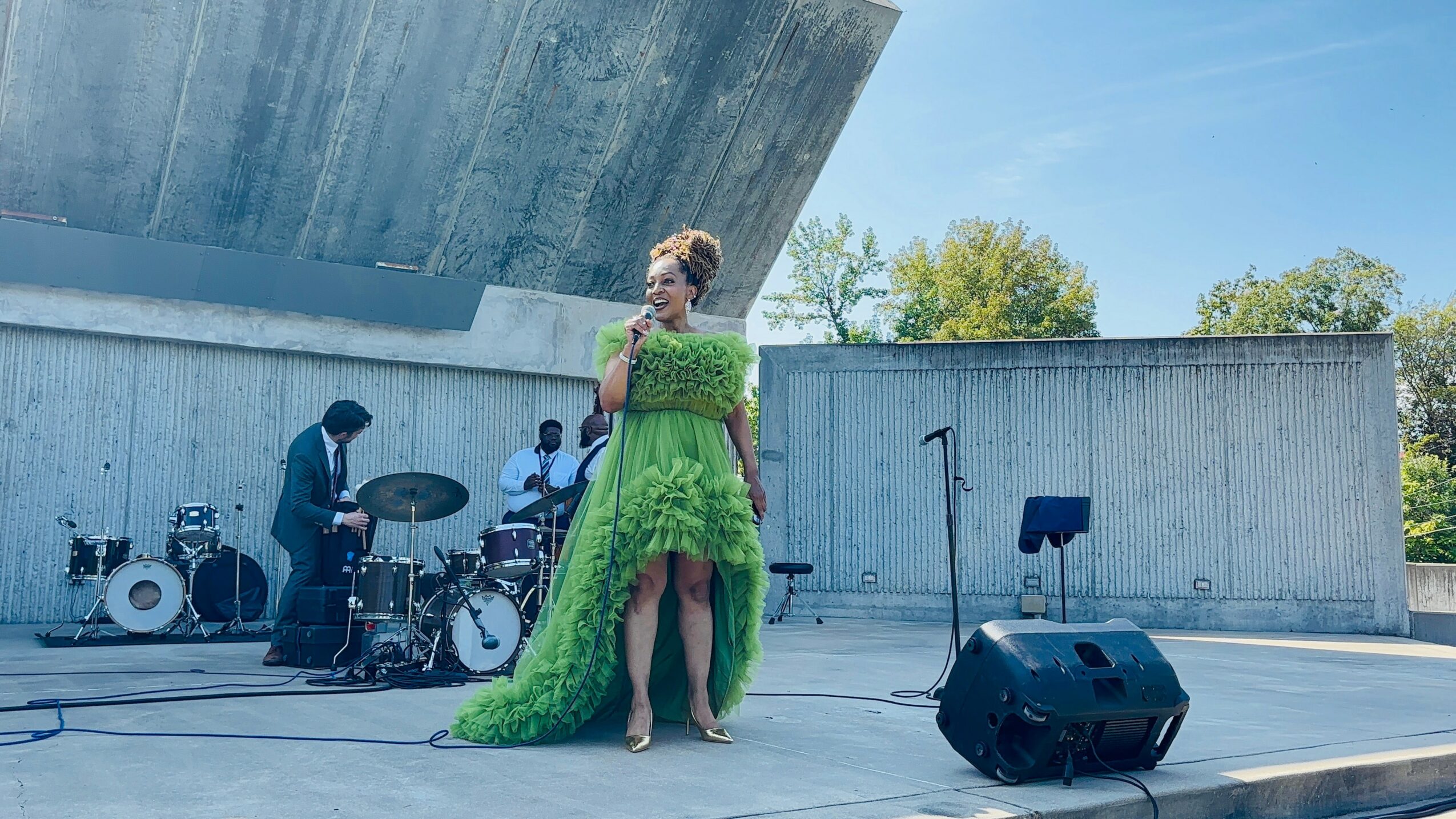 A woman in a green dress sings on an outdoor stage with musicians and drums, under a modern concrete structure.