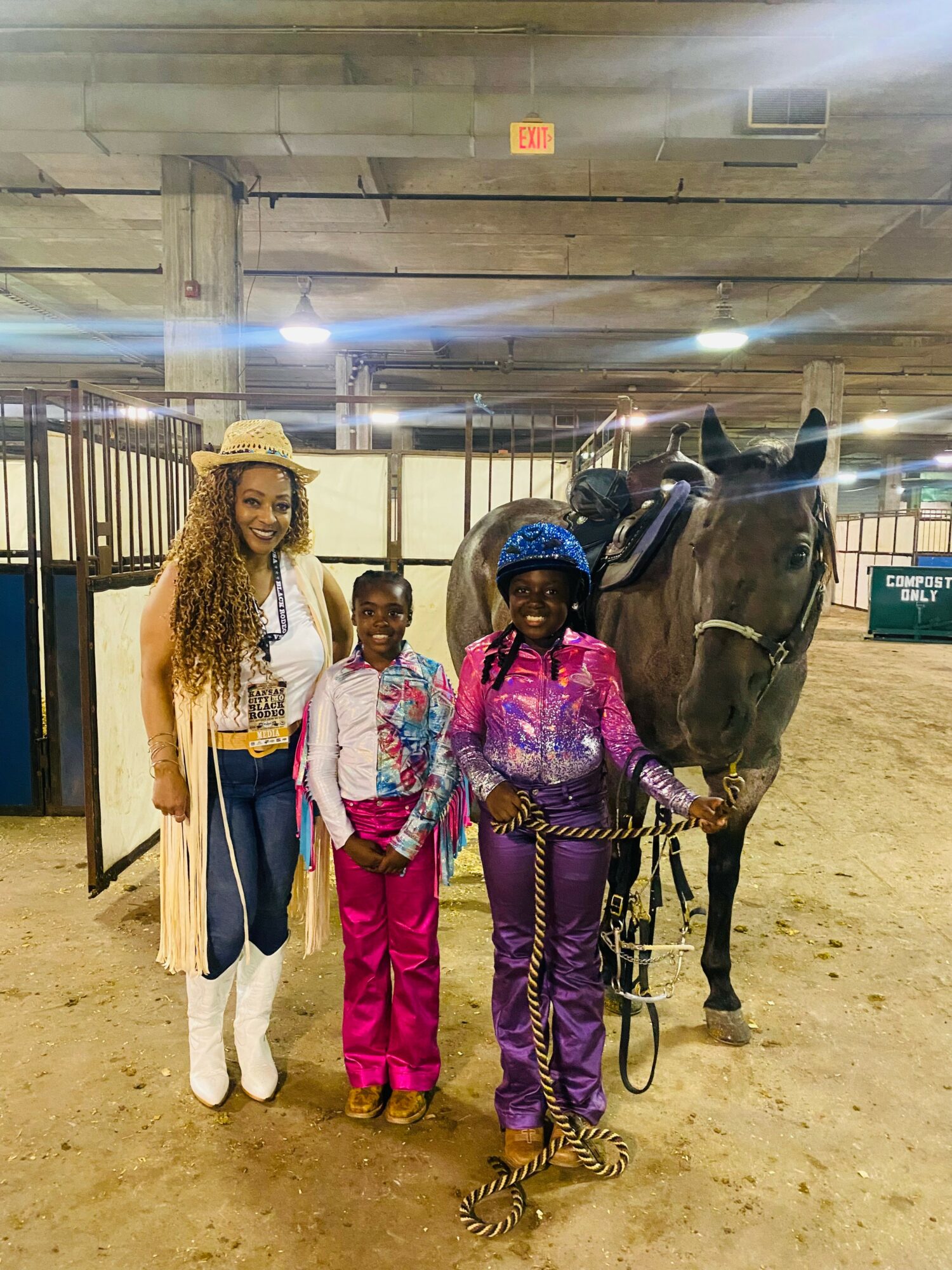 Three people and a horse in an indoor riding arena, with a woman on the left wearing a hat and two children in colorful clothing, one holding the horse's reins.